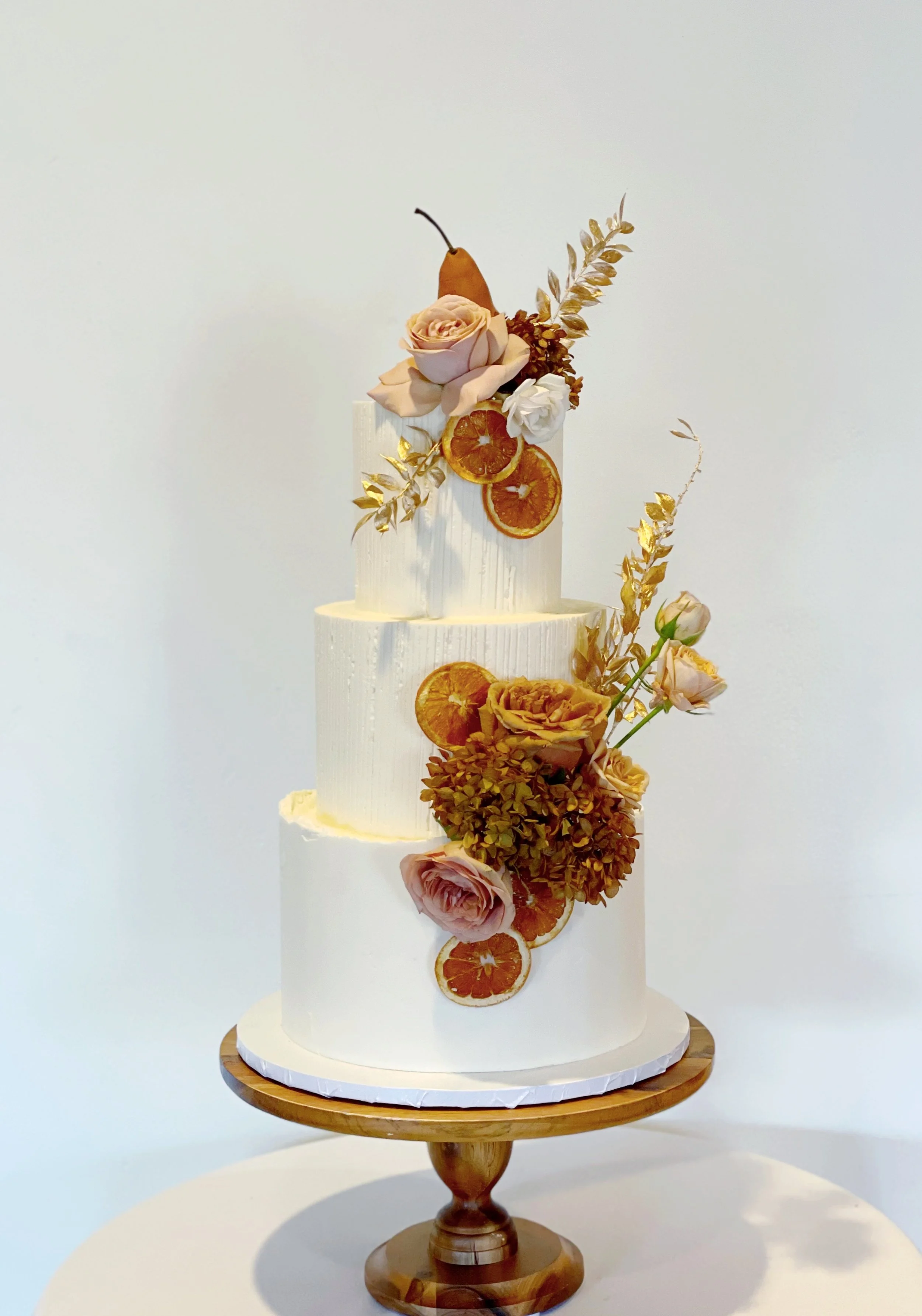 A three-tiered white wedding cake decorated with dried oranges, roses, and gold leaves on a wooden cake stand.