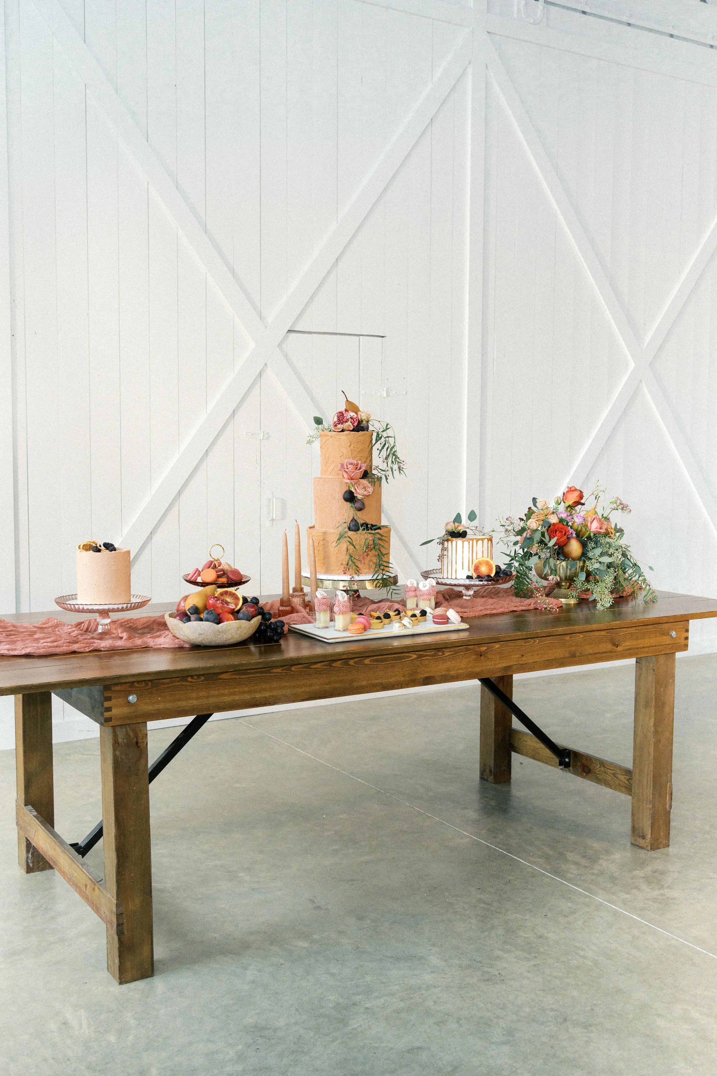 Decorative dessert table with a multi-tiered cake, assorted pastries, fresh fruit, and floral arrangements in a bright, modern space.