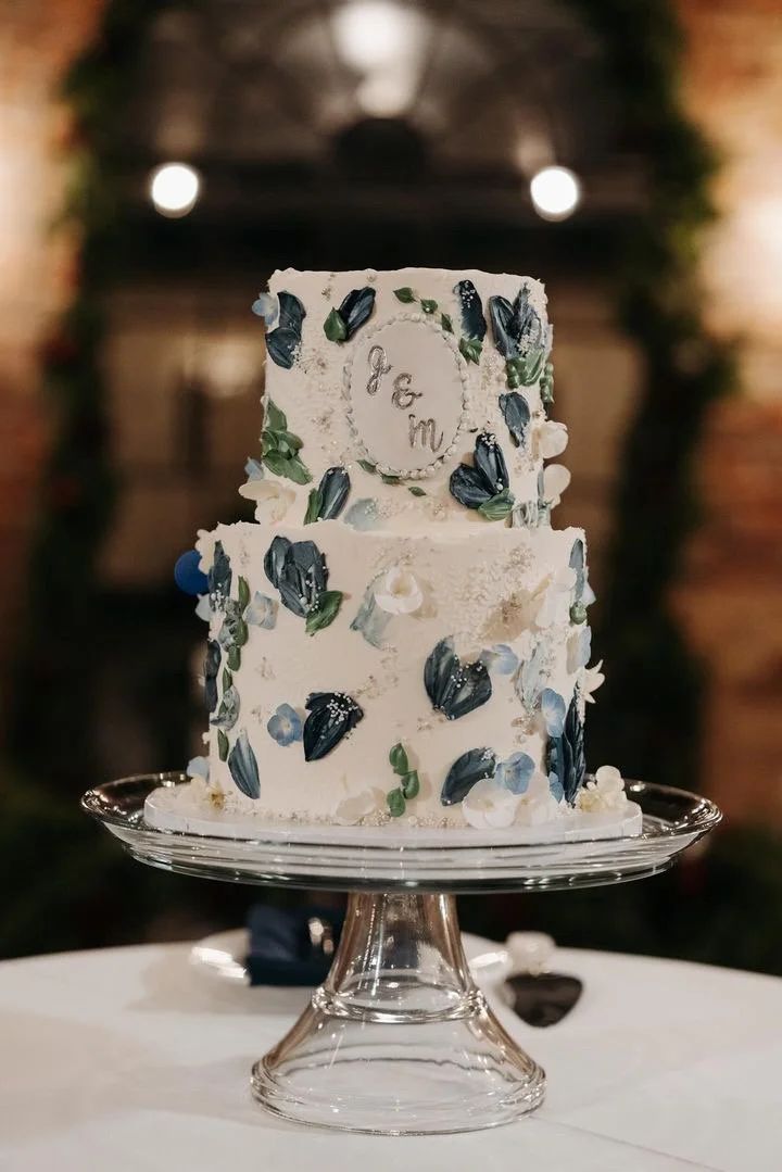Two-tier white wedding cake decorated with navy blue, green, and white flower and leaf accents, placed on a glass cake stand.