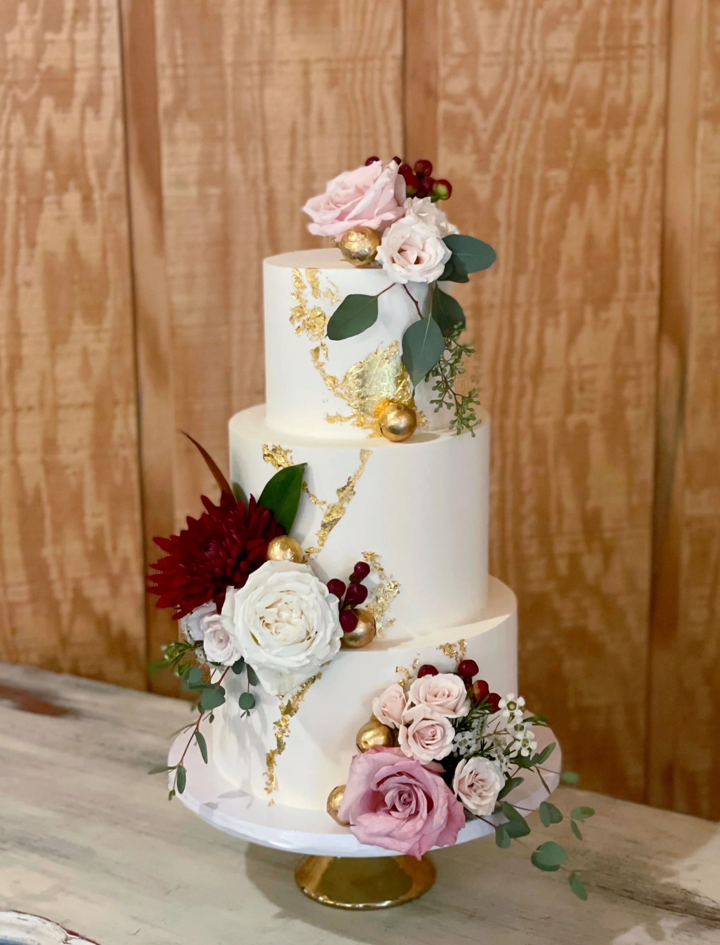 Three-tiered white wedding cake decorated with pink and white roses, red flowers, greenery, and gold accents placed on a gold cake stand.