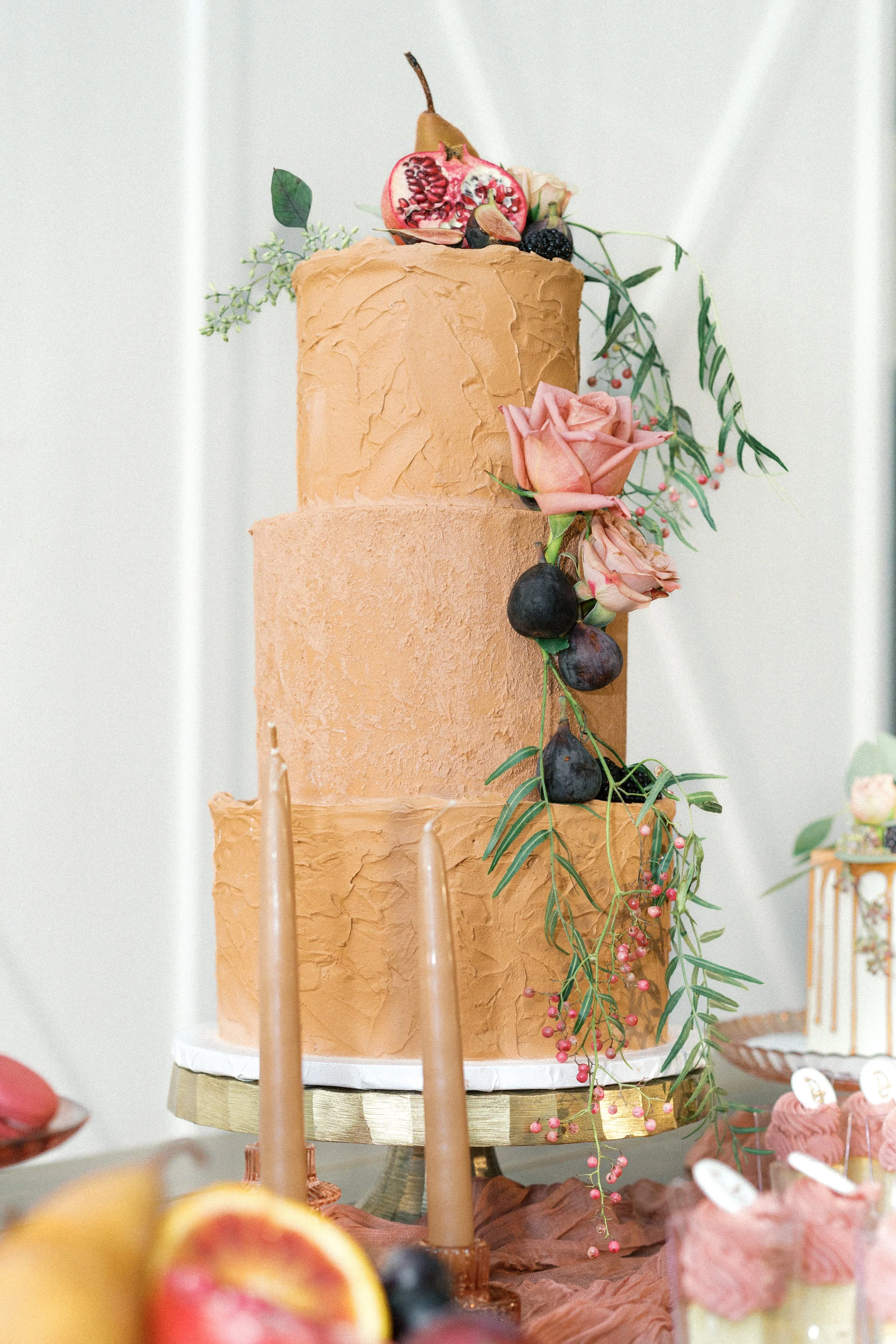 A three-tier peach-colored wedding cake decorated with pink roses, figs, blackberries, and greenery, placed on a gold cake stand.