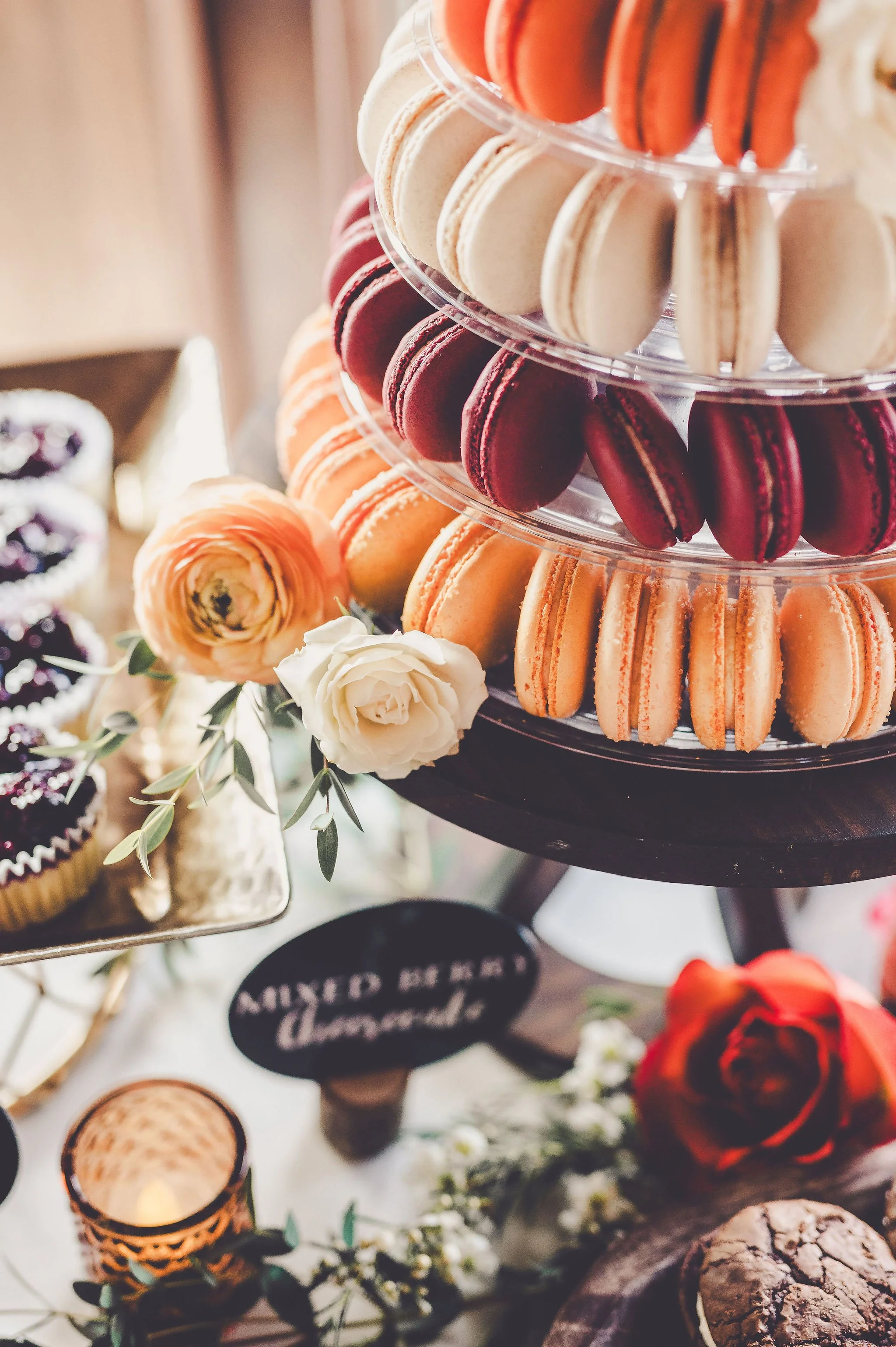A tiered stand filled with colorful macarons in orange, red, and beige, decorated with flowers, at a dessert table.