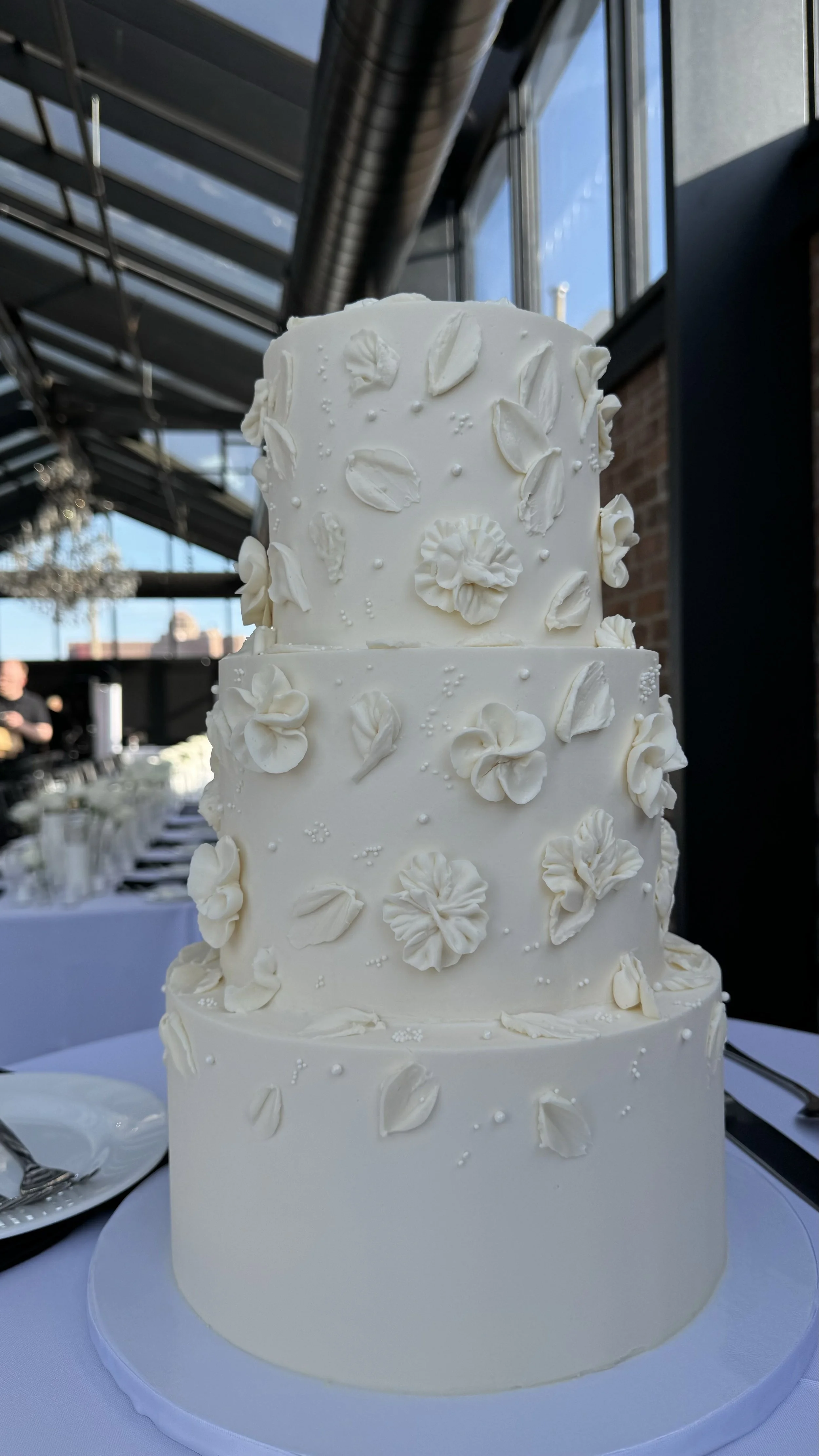 A three-tier white wedding cake decorated with white flower-shaped and petal decorations, placed on a white cake board on a white tablecloth table. The background shows a modern building interior with large glass windows and black metal beams.