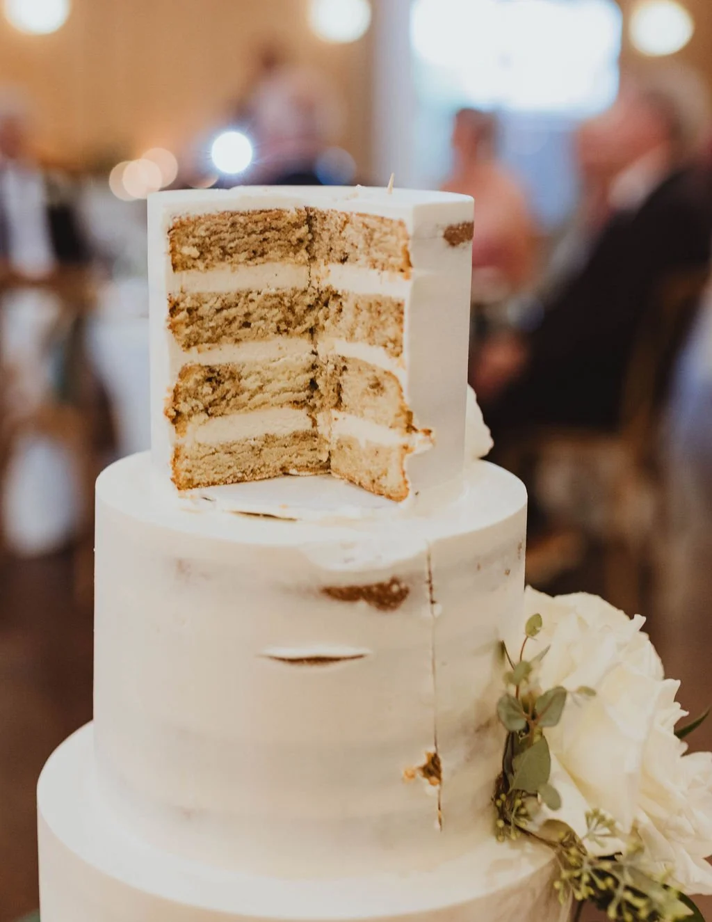A white, tiered wedding cake with a slice removed, revealing golden sponge layers and cream filling, decorated with white flowers and green foliage, in a blurred reception hall.