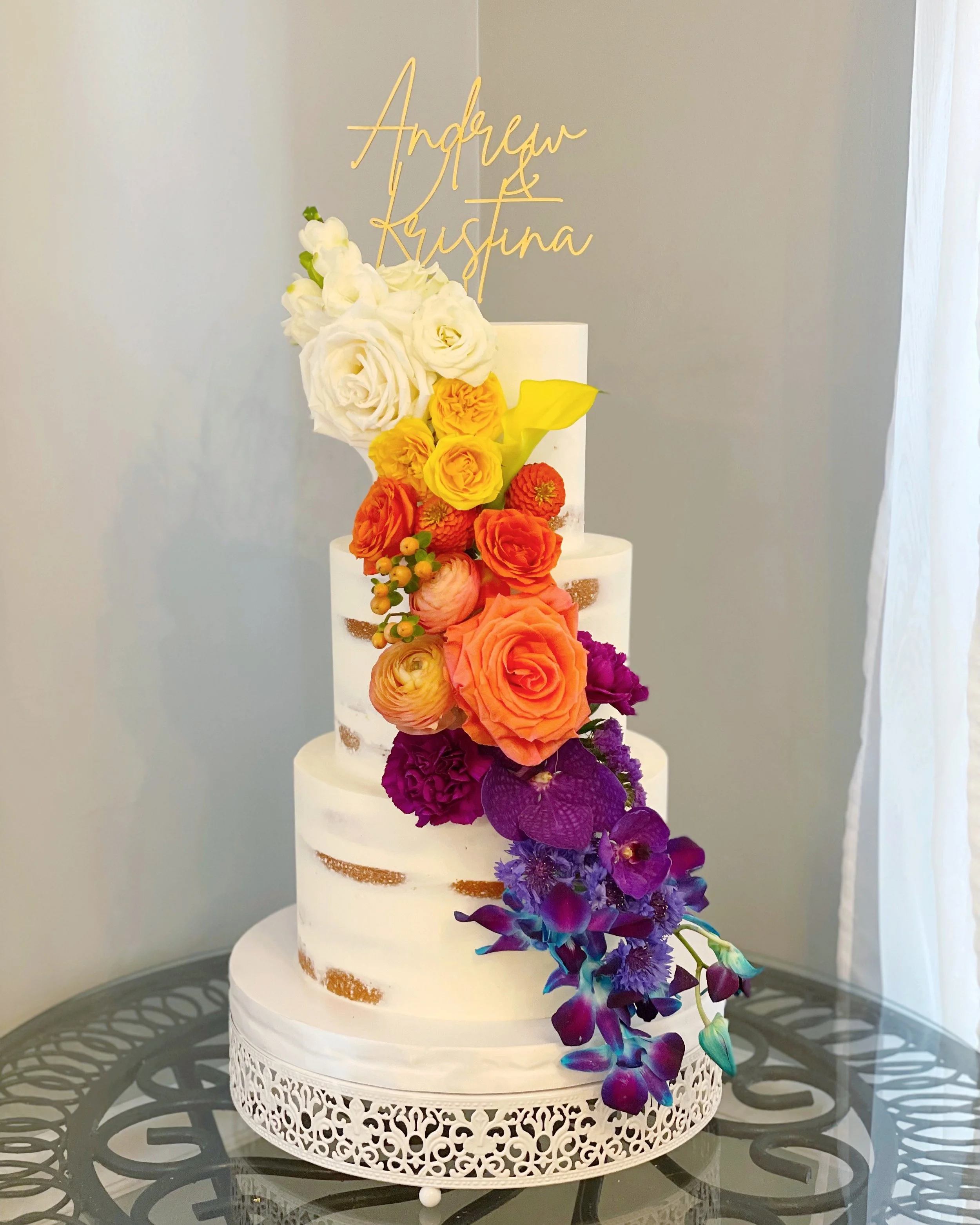 A tall, multi-tiered white wedding cake decorated with colorful flowers and a gold-colored cake topper with the names 'Andrew & Kristina'.