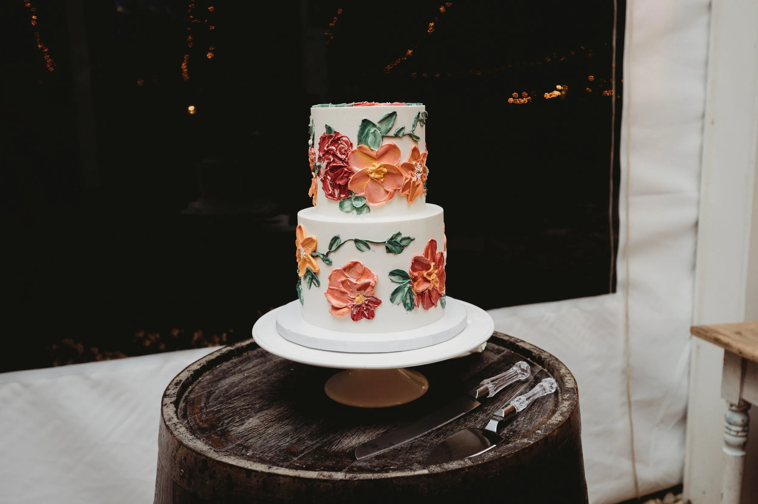 Two-tiered white wedding cake decorated with colorful flowers, placed on a white cake stand on a wooden table with cake serving utensils nearby.