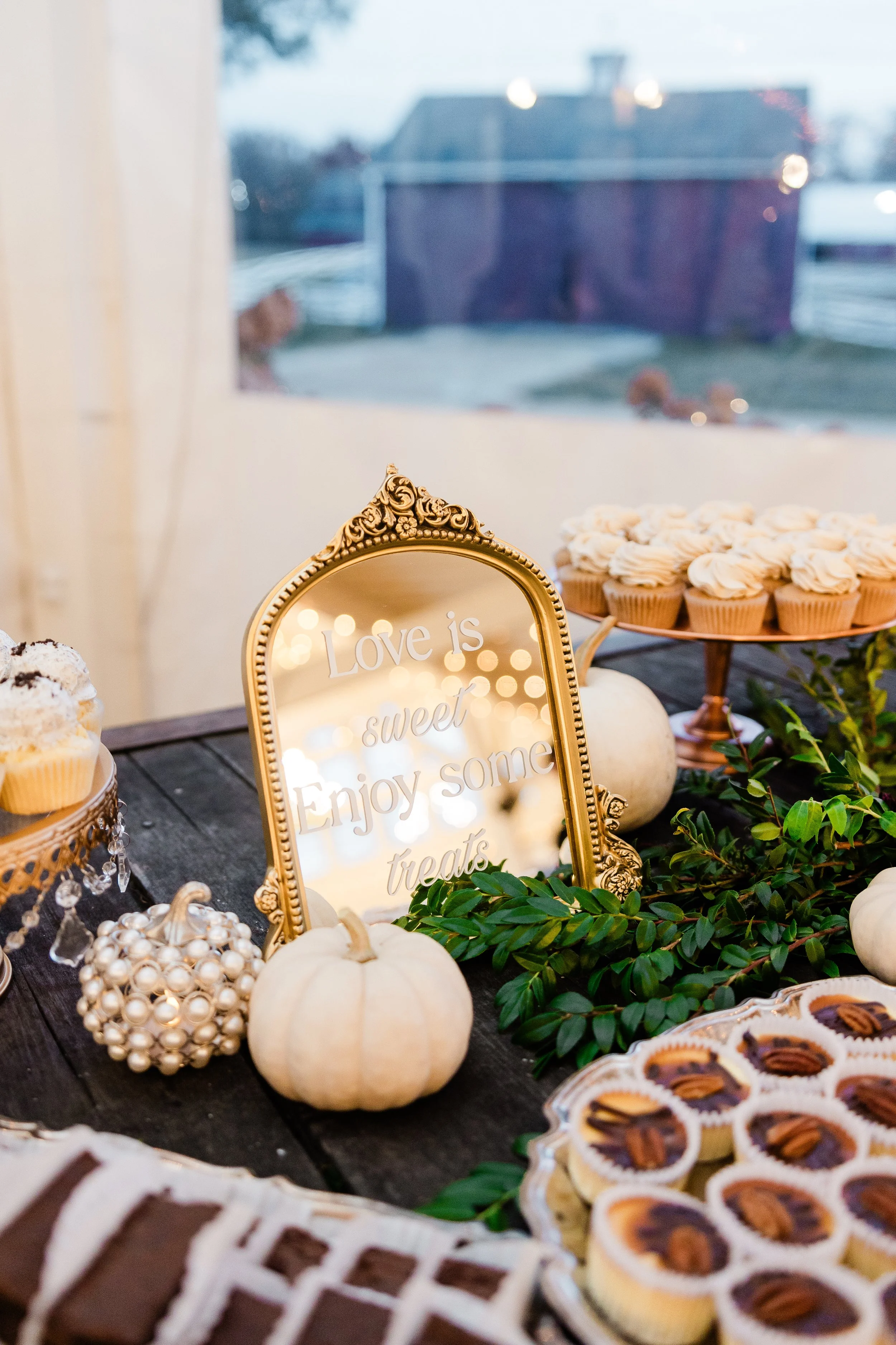 Decorative dessert table with white pumpkins, cupcakes, and a mirror with the phrase "Love is sweet, enjoy some treats." The table is adorned with greenery and is set near a window with a blurry outdoor scene in the background.
