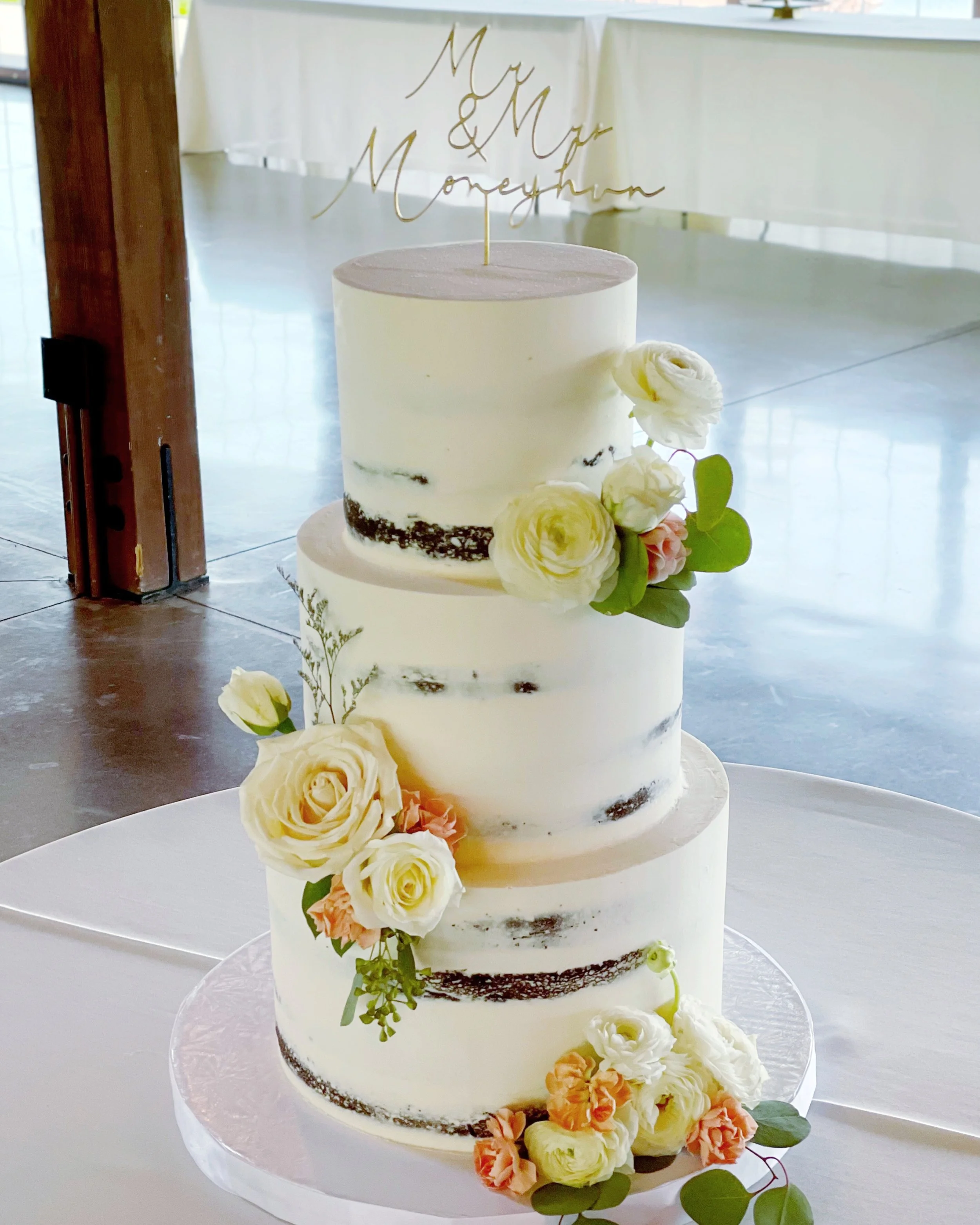 A three-tiered wedding cake with white buttercream, decorated with fresh cream-colored and blush pink flowers and green leaves, topped with a gold topper that reads 'Mr & Mrs Moneymon'.
