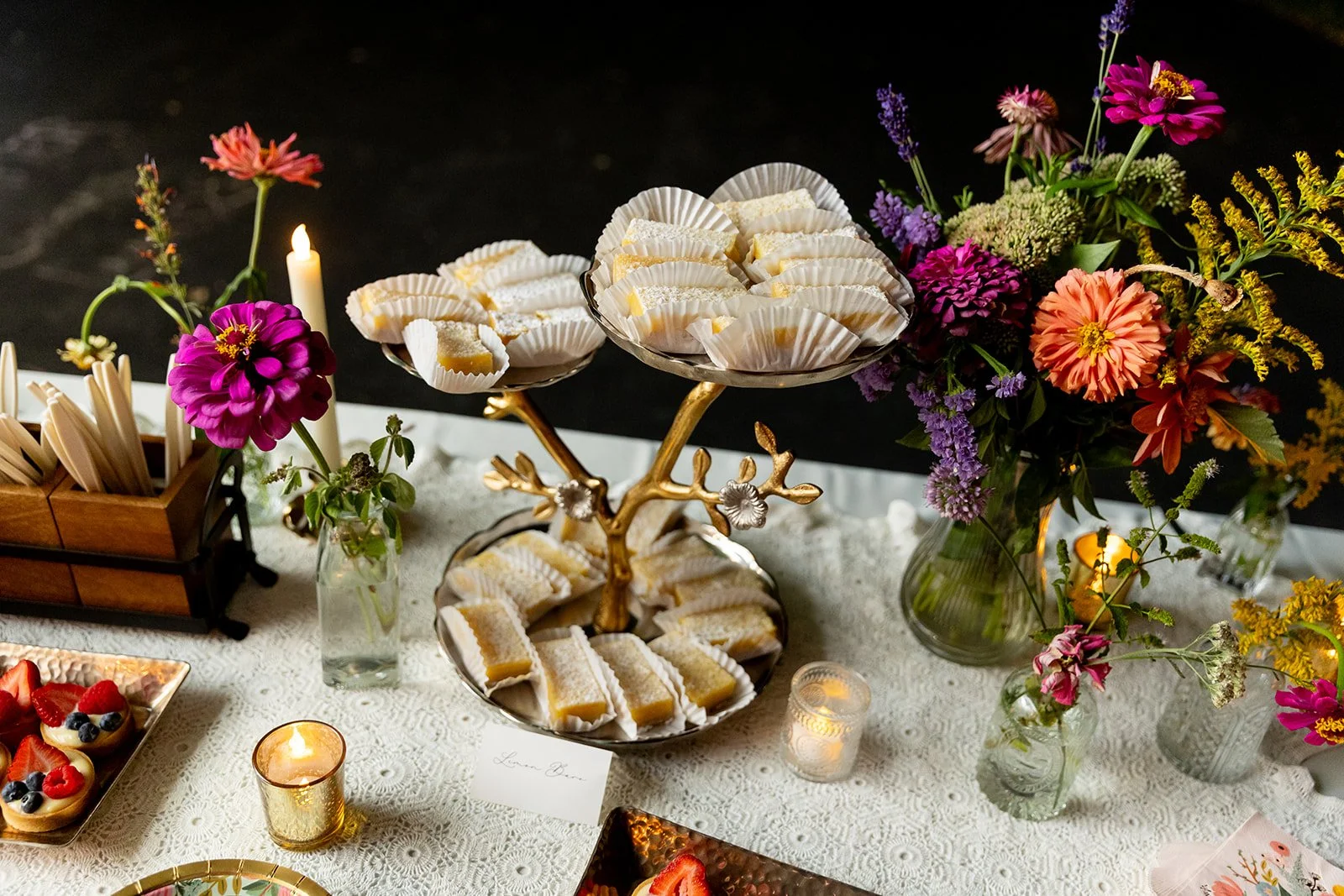 Dessert table with floral arrangements, featuring trays of lemon bars with powdered sugar, candles, and small fruit tarts, set on a white lace tablecloth.