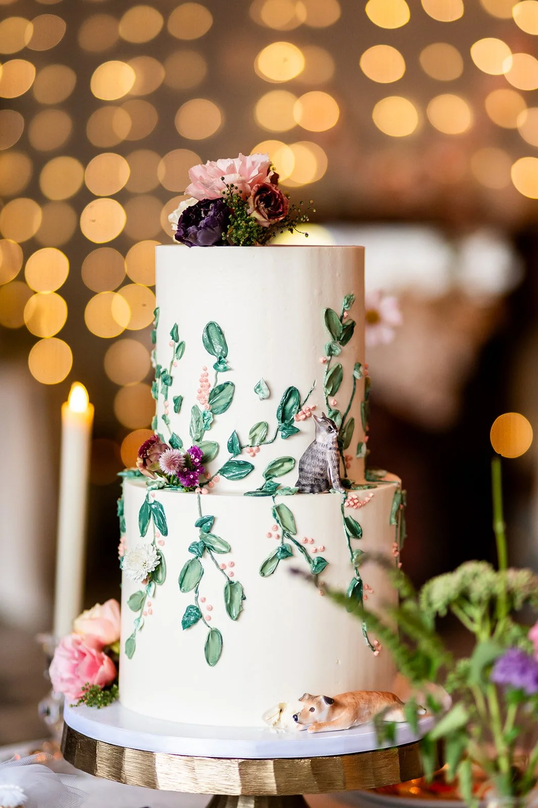 A tall, white, tiered wedding cake decorated with green leaves, pink berries, and floral accents, topped with purple and pink flowers. There are small dog figurines at the bottom and middle layers, and a background with warm, blurred lights.