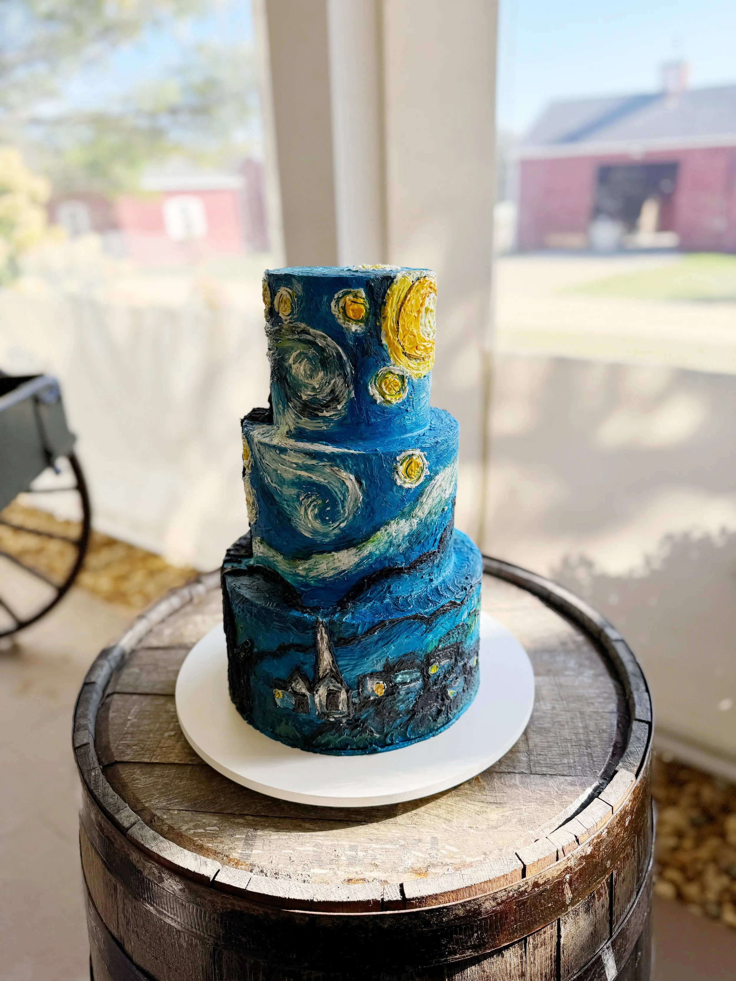 A three-tiered cake inspired by Vincent van Gogh's Starry Night painting, featuring swirling night sky, bright stars, a crescent moon, and a village with houses and a church, placed on a white cake board on a wooden barrel.