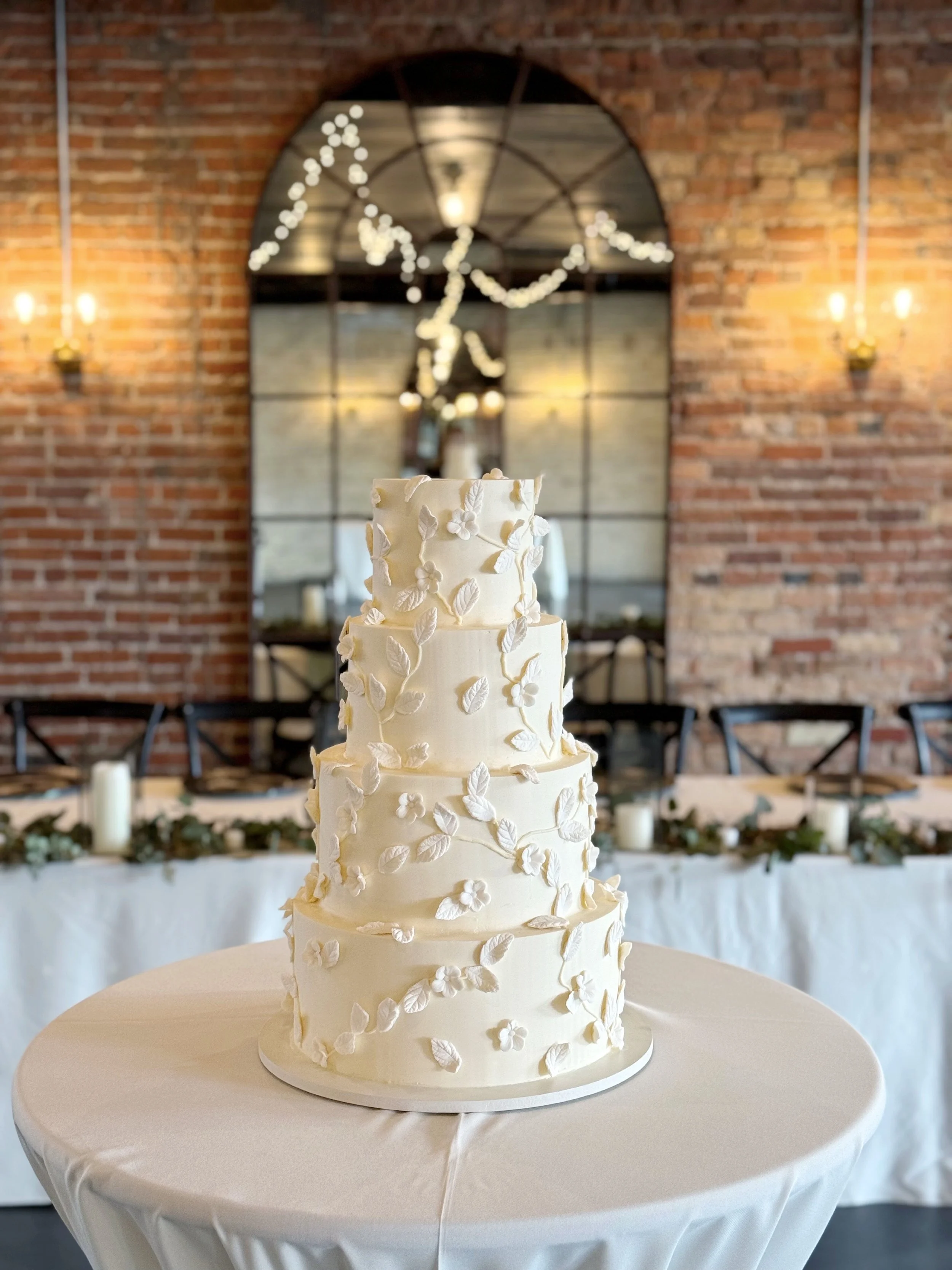 A multi-tiered white wedding cake decorated with white floral and leaf patterns, placed on a round table covered with a white tablecloth in a rustic wedding venue with exposed brick walls and a large mirror in the background.
