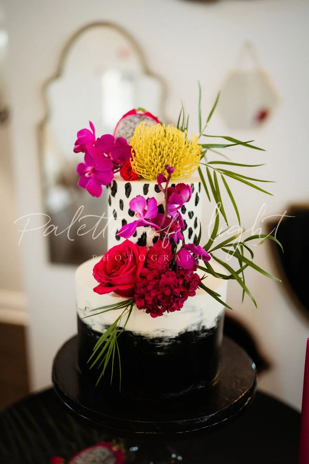 A modern black and white layered cake decorated with vibrant pink, yellow, and red flowers, with peacock feather details, placed on a black cake stand.