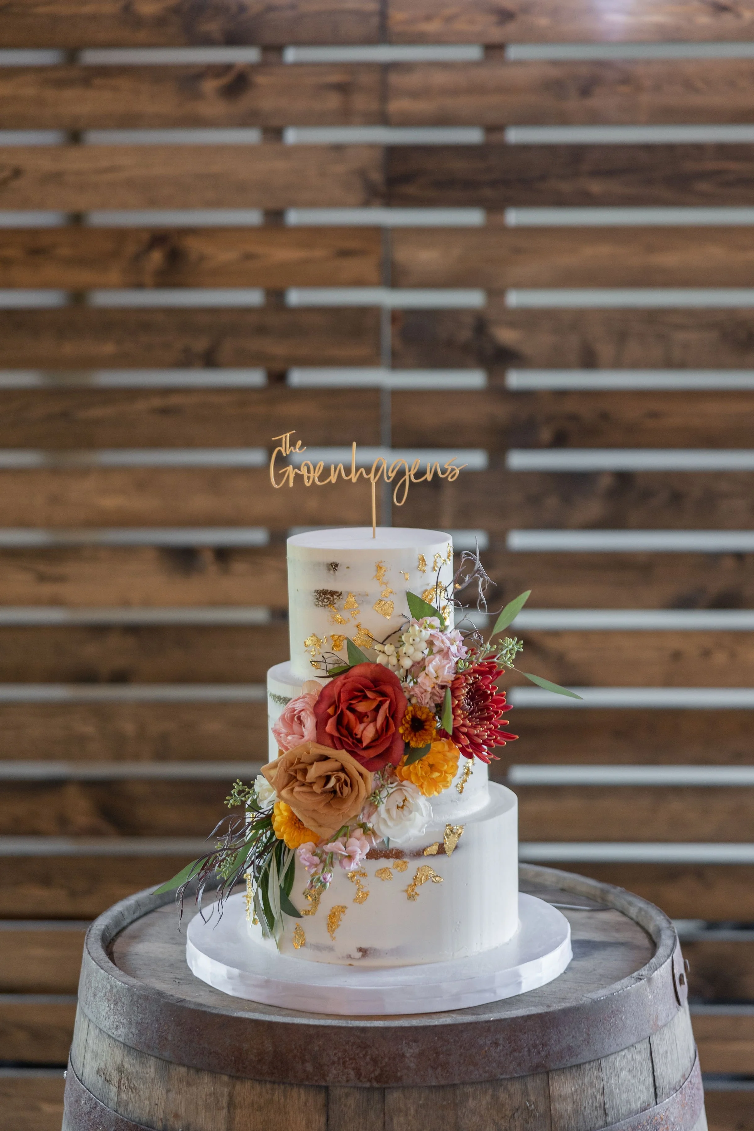 A three-tier white wedding cake decorated with colorful flowers and gold accents, topped with a gold cake topper reading 'The Groenhagens', placed on a wooden barrel against a wooden backdrop.