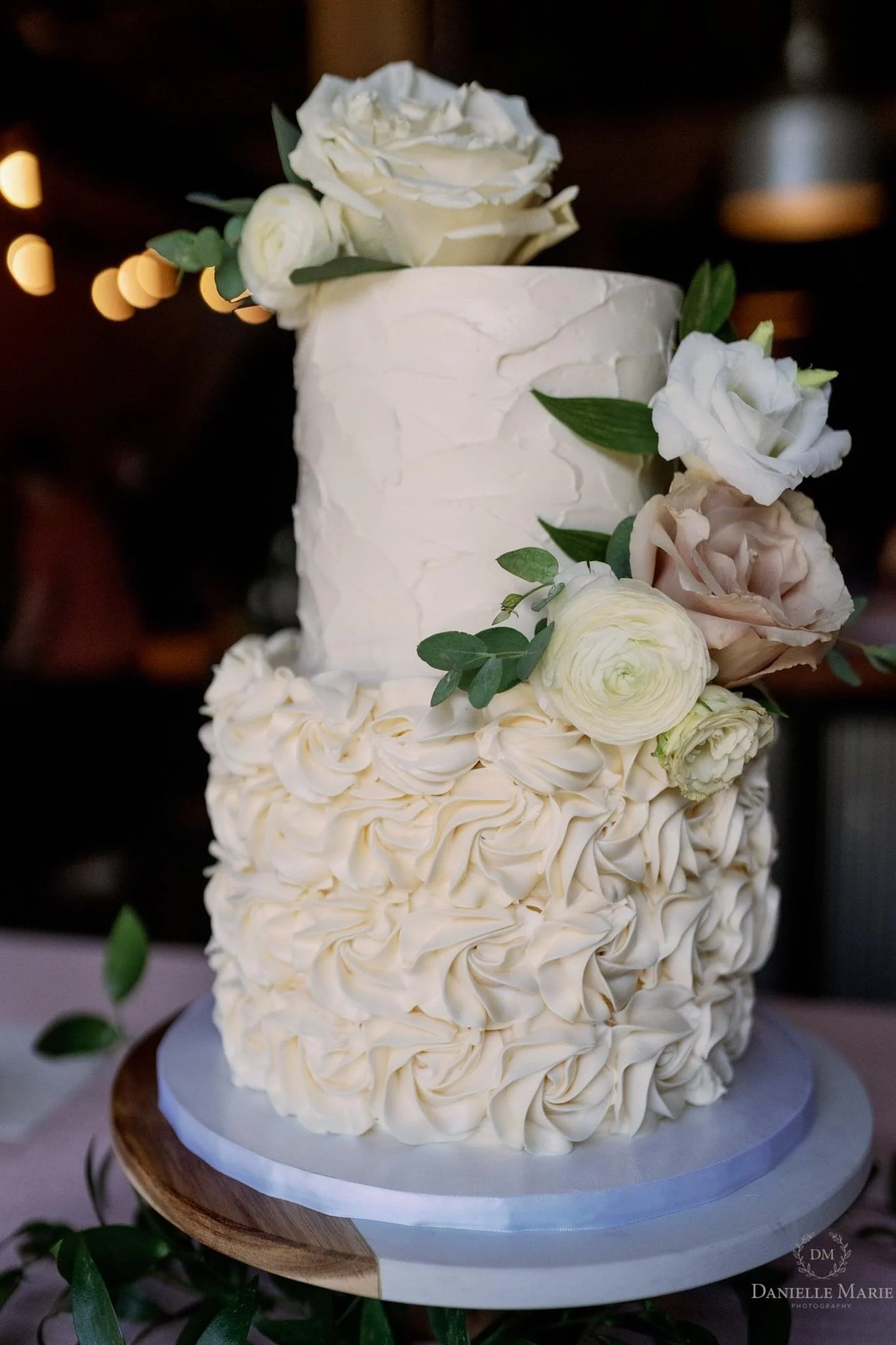 A two-tiered wedding cake decorated with white and cream-colored flowers and green leaves, with textured icing and rosette decorations.