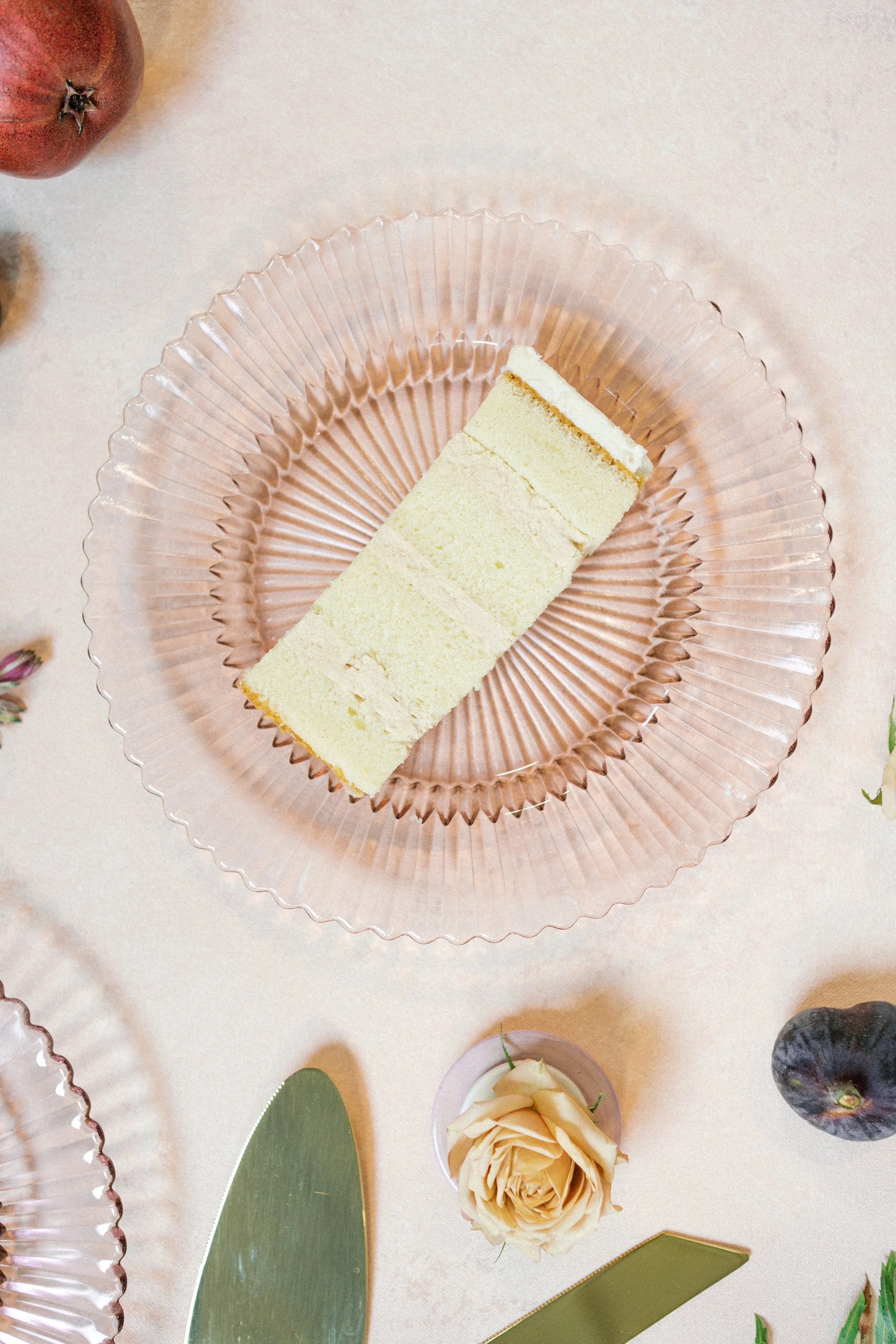 A piece of layered cake on a glass plate surrounded by a flower, a fig, and a couple of green leaves on a cream-colored tablecloth.