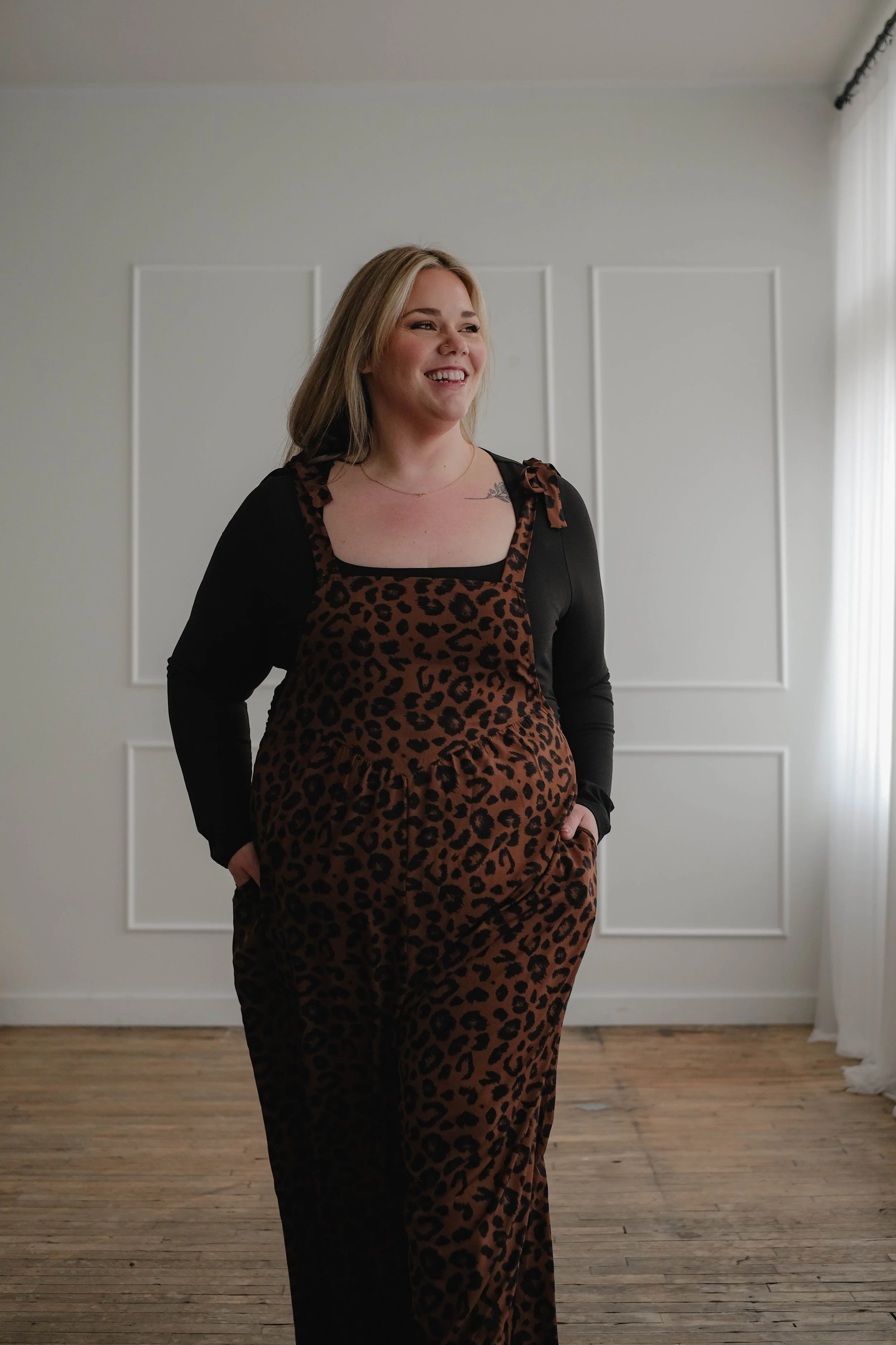 A woman with blonde hair smiling, standing indoors wearing a black long-sleeve top and brown leopard print overalls.