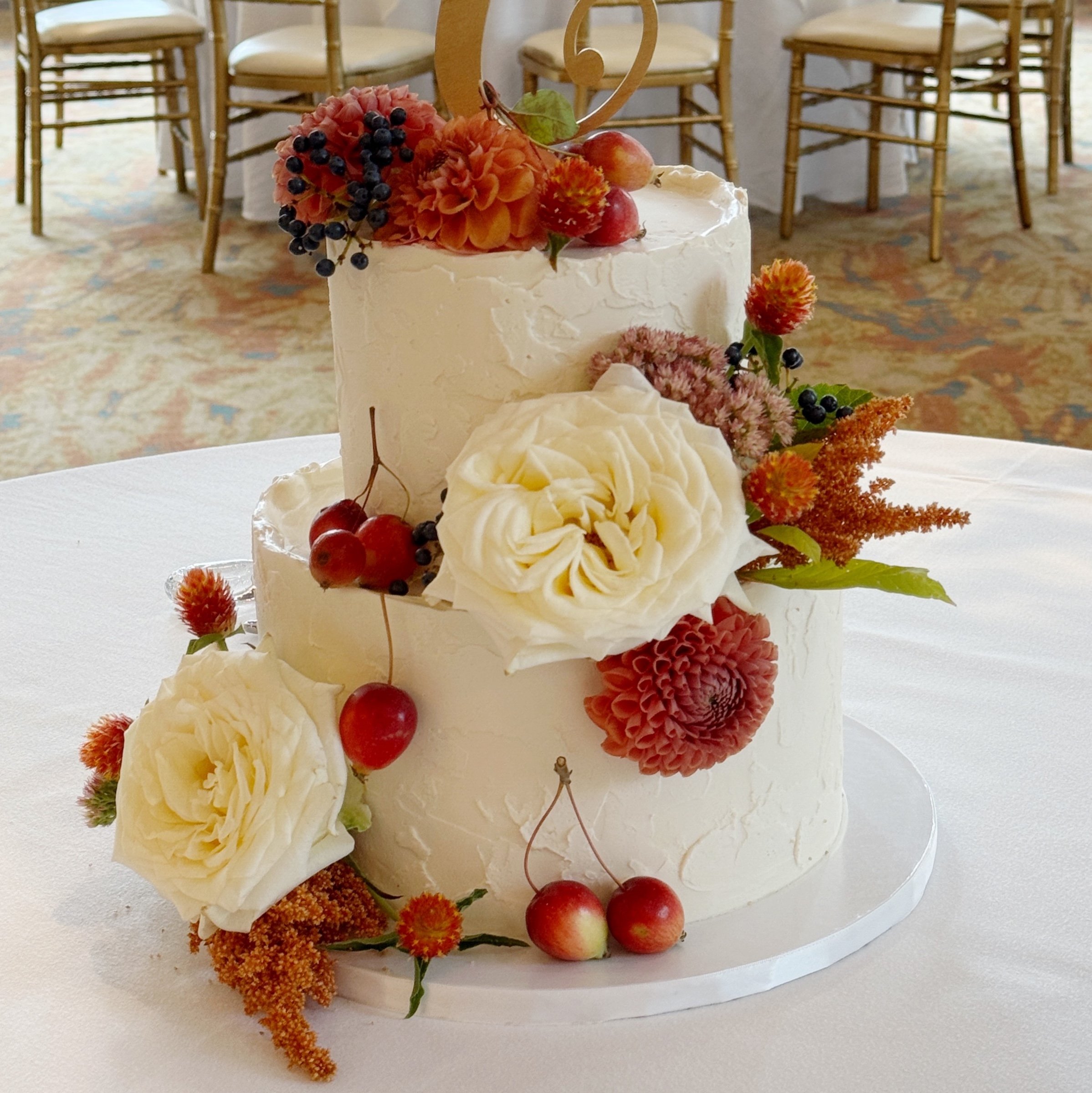 Two-tiered wedding cake decorated with white and red flowers, red cherries, black berries, and greenery, with a gold number "2" cake topper, placed on a white tablecloth at an indoor event with gold chairs in the background.
