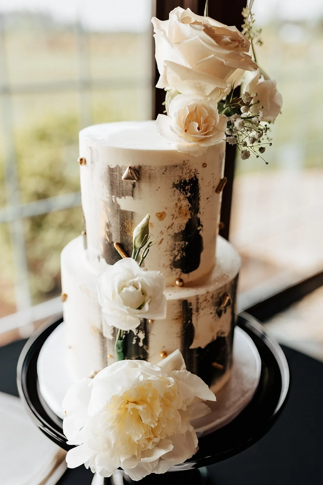 A two-tiered white wedding cake decorated with white roses, greenery, and gold accents, placed on a black cake stand near a window.