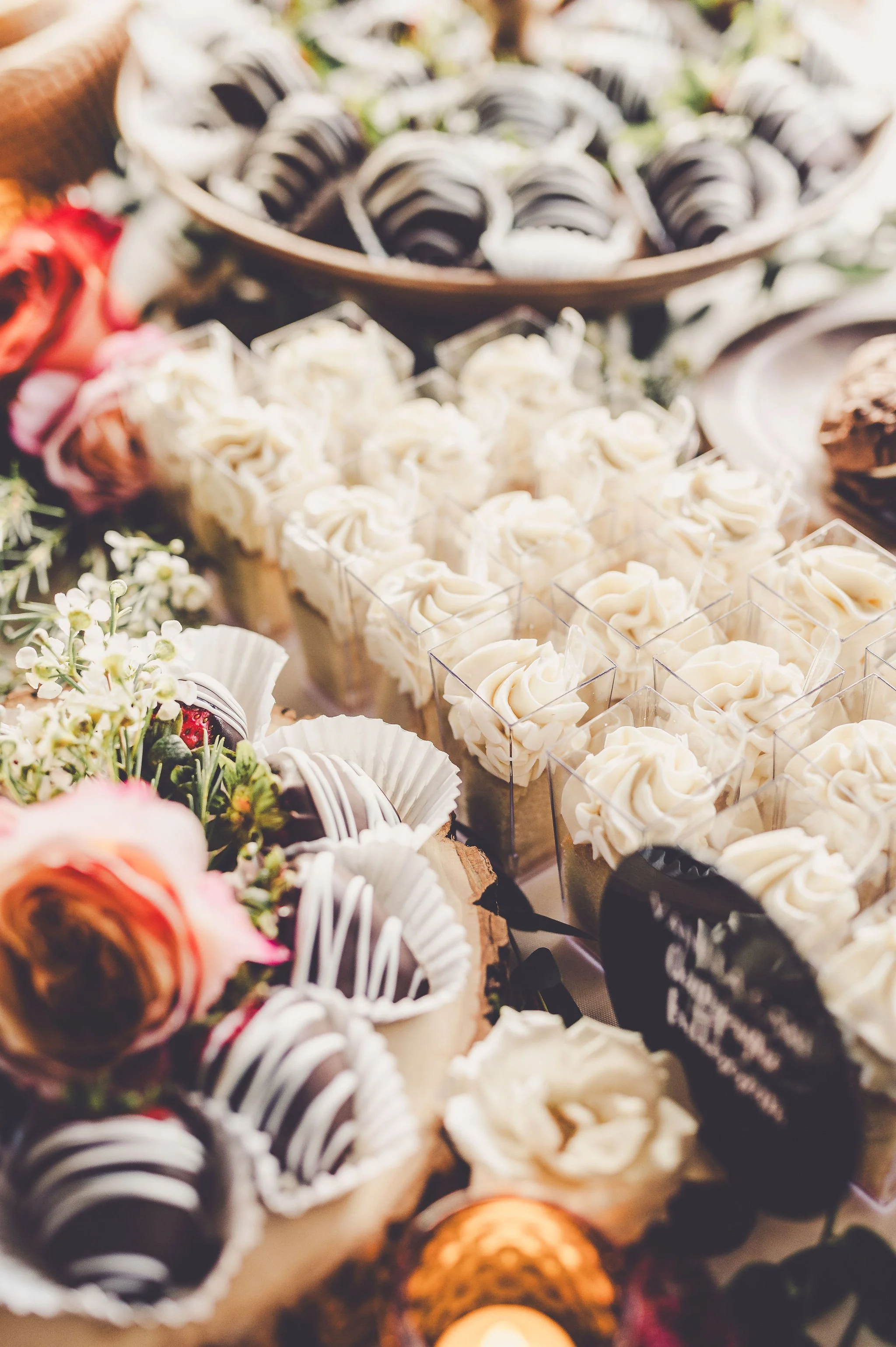 Assorted cupcakes and small desserts on a decorated table, topped with frosting, flowers, and chocolate.