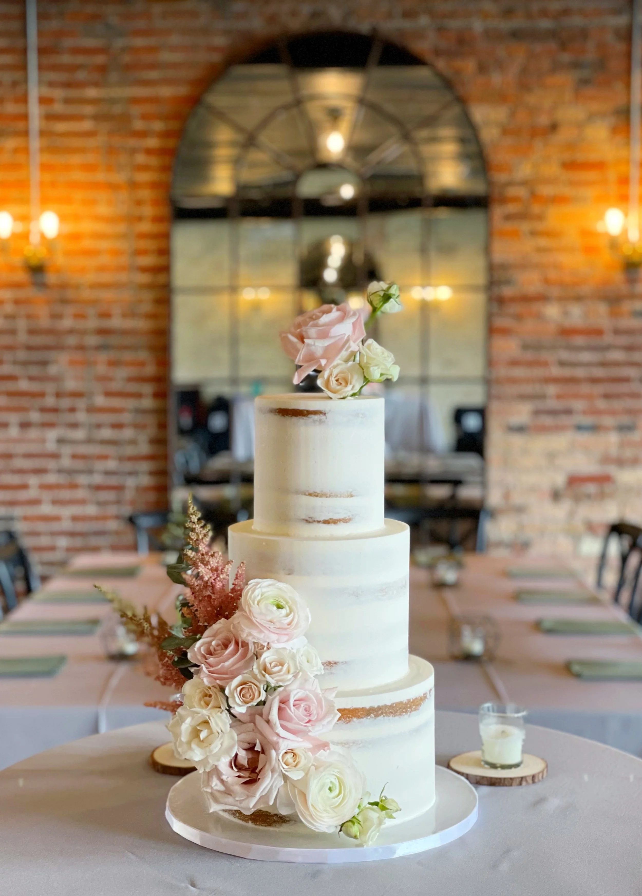 A three-tier wedding cake with white frosting, decorated with pale pink and white roses and greenery, in a rustic venue with brick walls and a large window mirror in the background.
