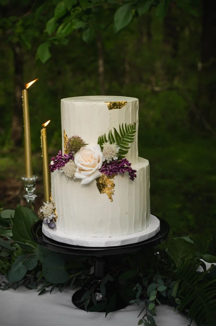 A two-tier white wedding cake decorated with fresh flowers and greenery, placed on a black cake stand with lit gold candles nearby, outdoors amidst green foliage.