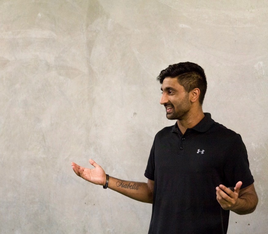 Man with dark hair, beard, wearing a black Under Armour polo shirt, smiling, standing against a plain, textured wall, gesturing with his right hand.