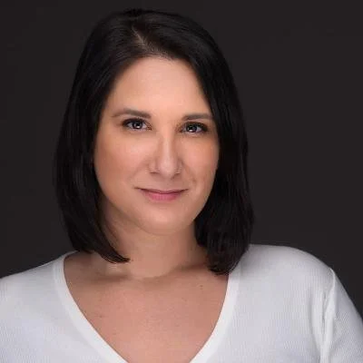 Headshot of a woman with shoulder-length dark hair, wearing a white top, smiling slightly against a dark gray background.