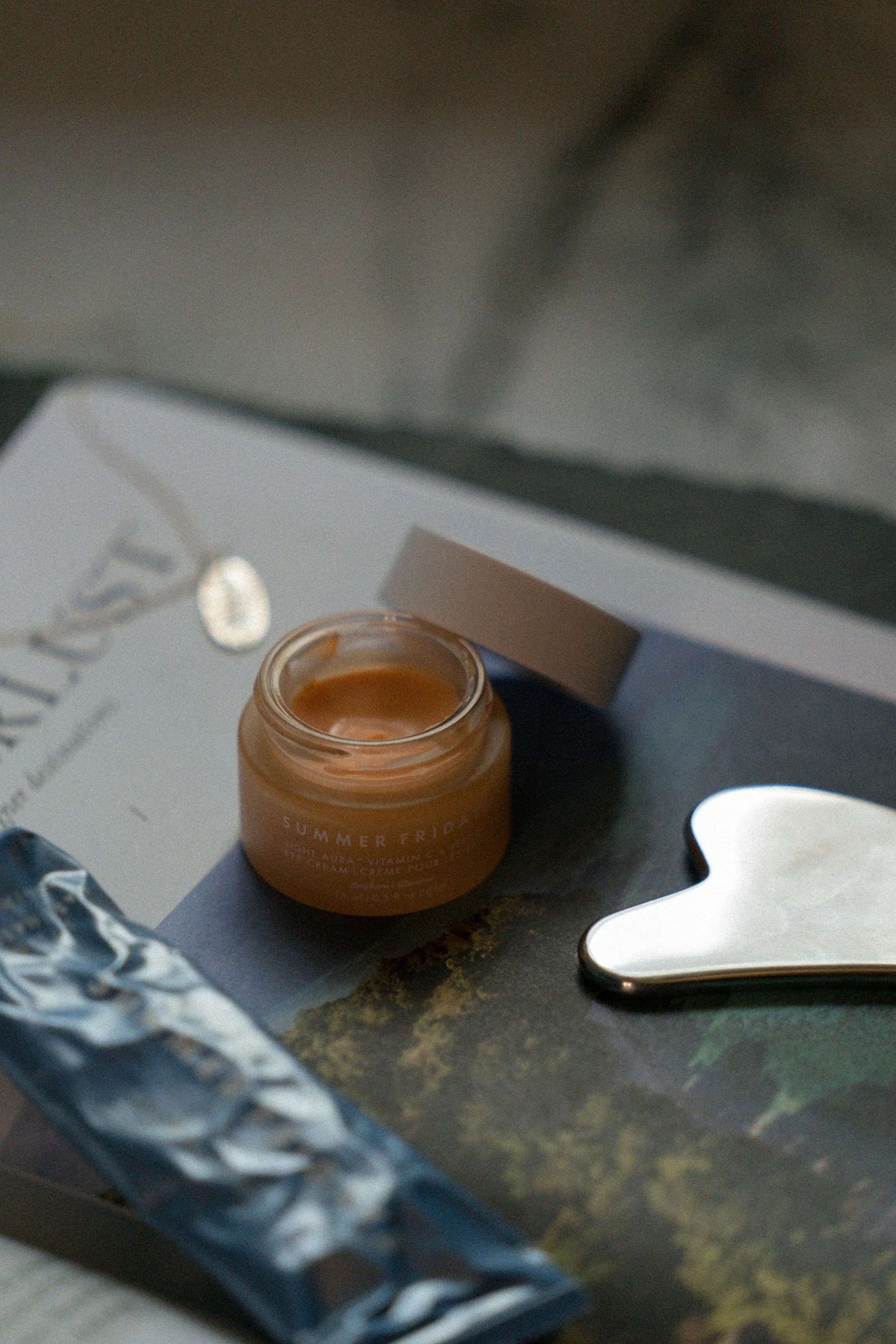 Open jar of Summer Fridays bright algae vitamin-dream cream with a white lid nearby, on a dark surface with a white paper or box and a silver spoon.