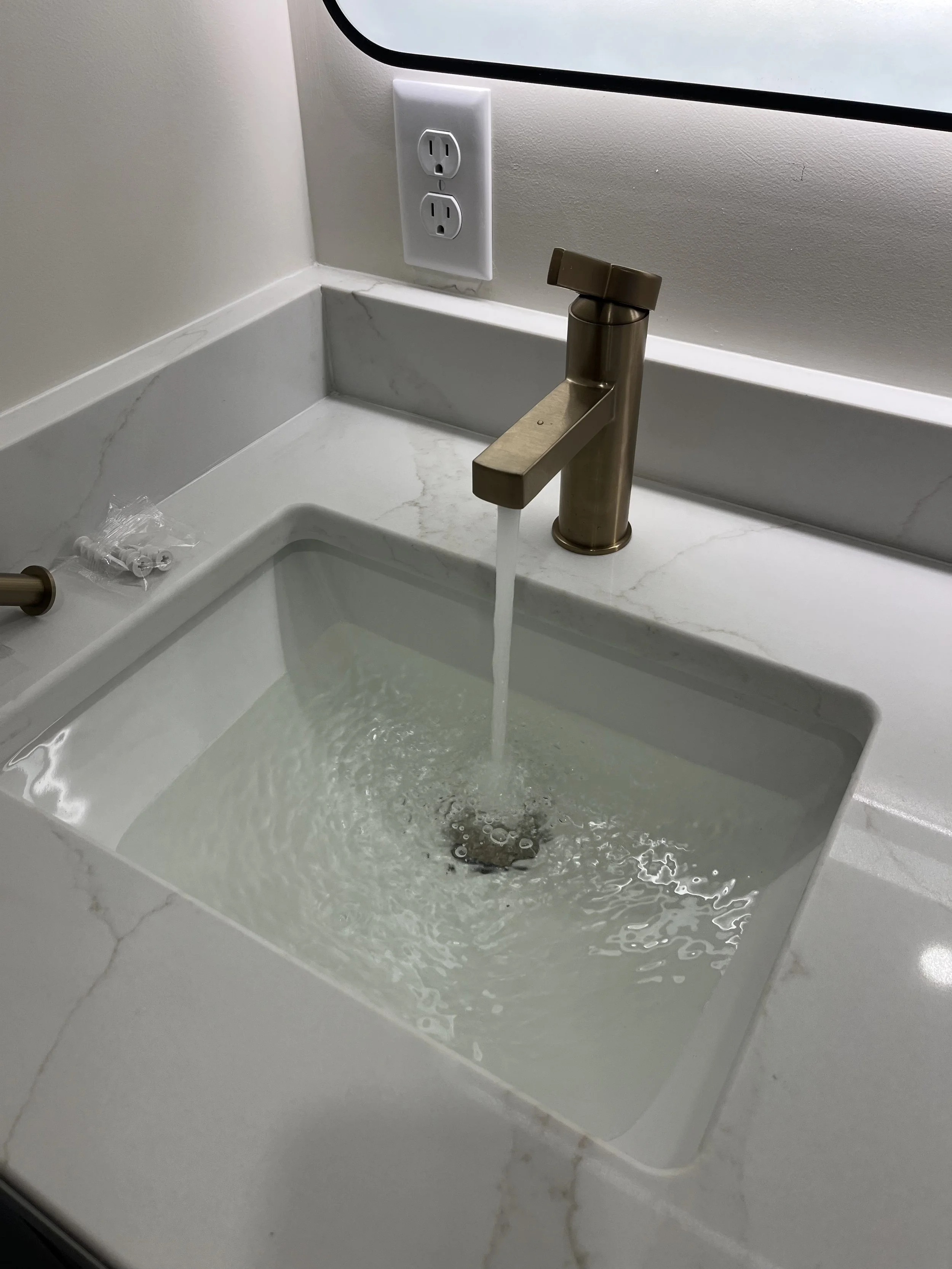 A kitchen countertop with a white marble pattern and a modern brass faucet dispensing water into a rectangular sink, with a wall outlet and a mirror above.
