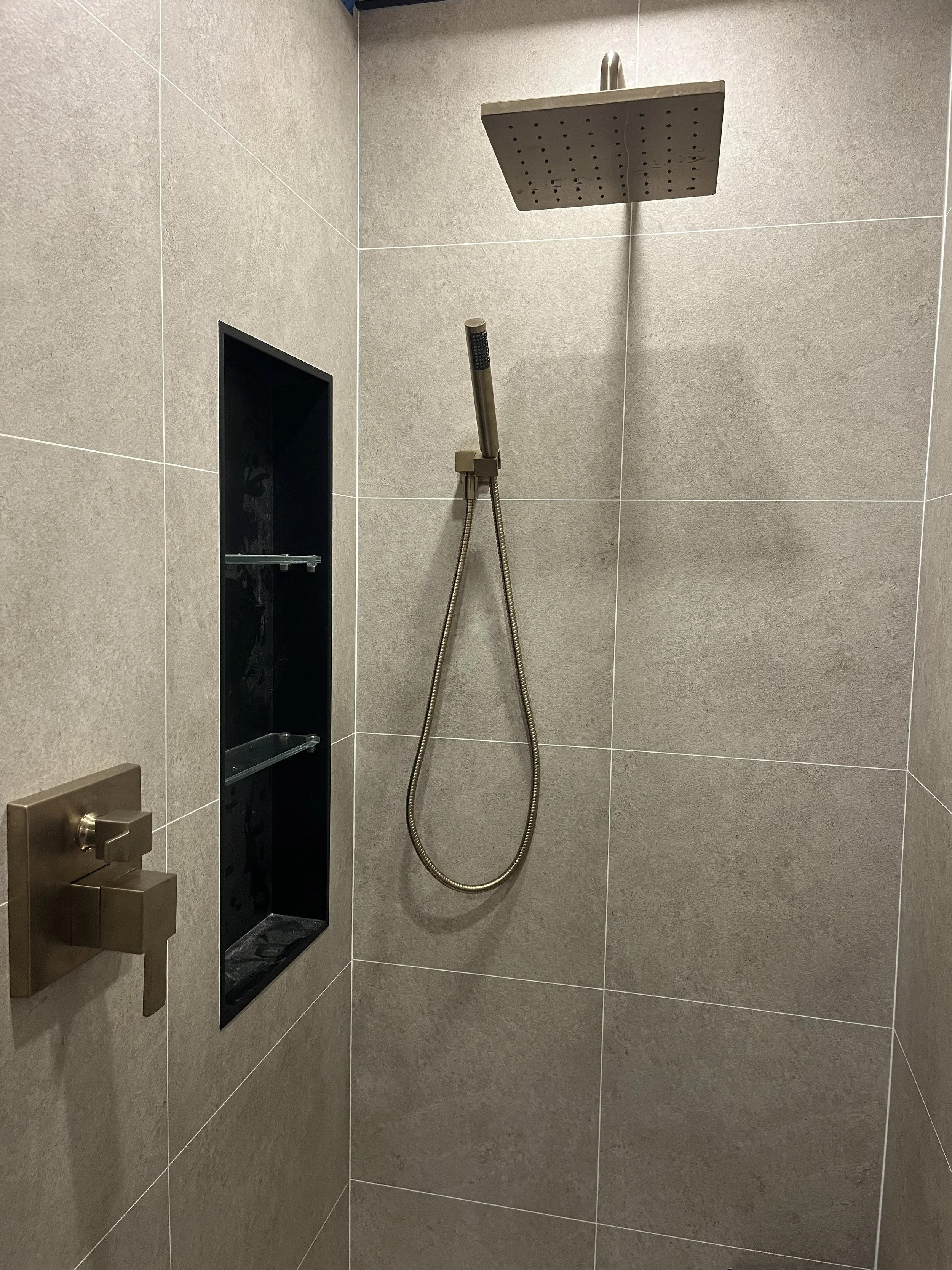 Shower area with a large square overhead showerhead, a handheld showerhead attached by a flexible hose, a built-in black shower niche, and a brushed bronze shower control handle.