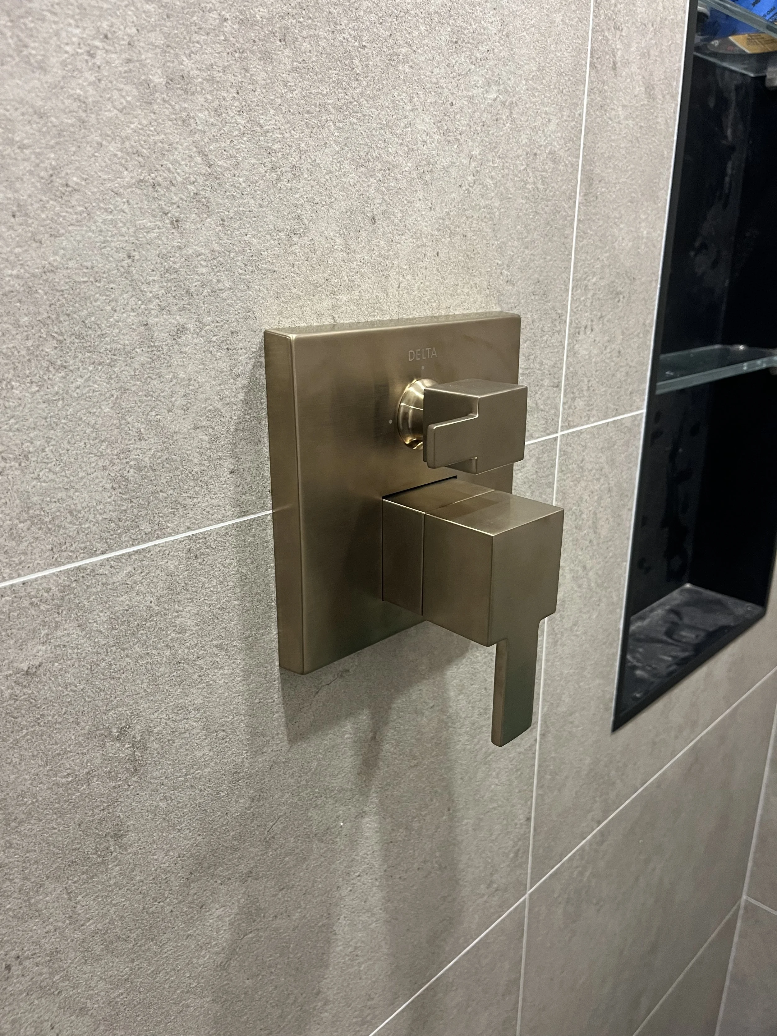 Close-up of a metallic flush valve handle on a tiled bathroom wall.