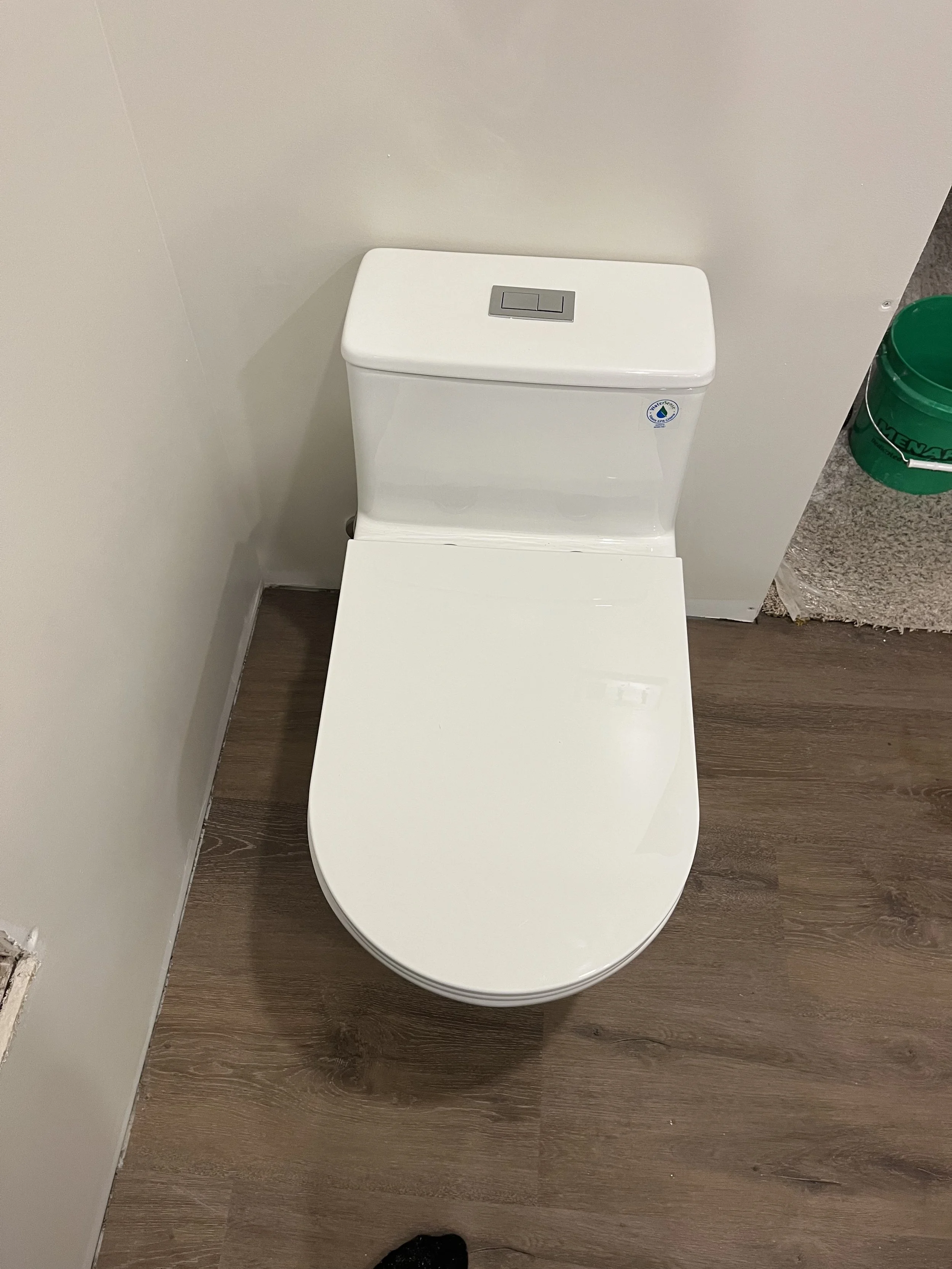 A white modern toilet with a rectangular flush button on the tank, installed against a light-colored wall, next to a green bucket on a wooden floor.