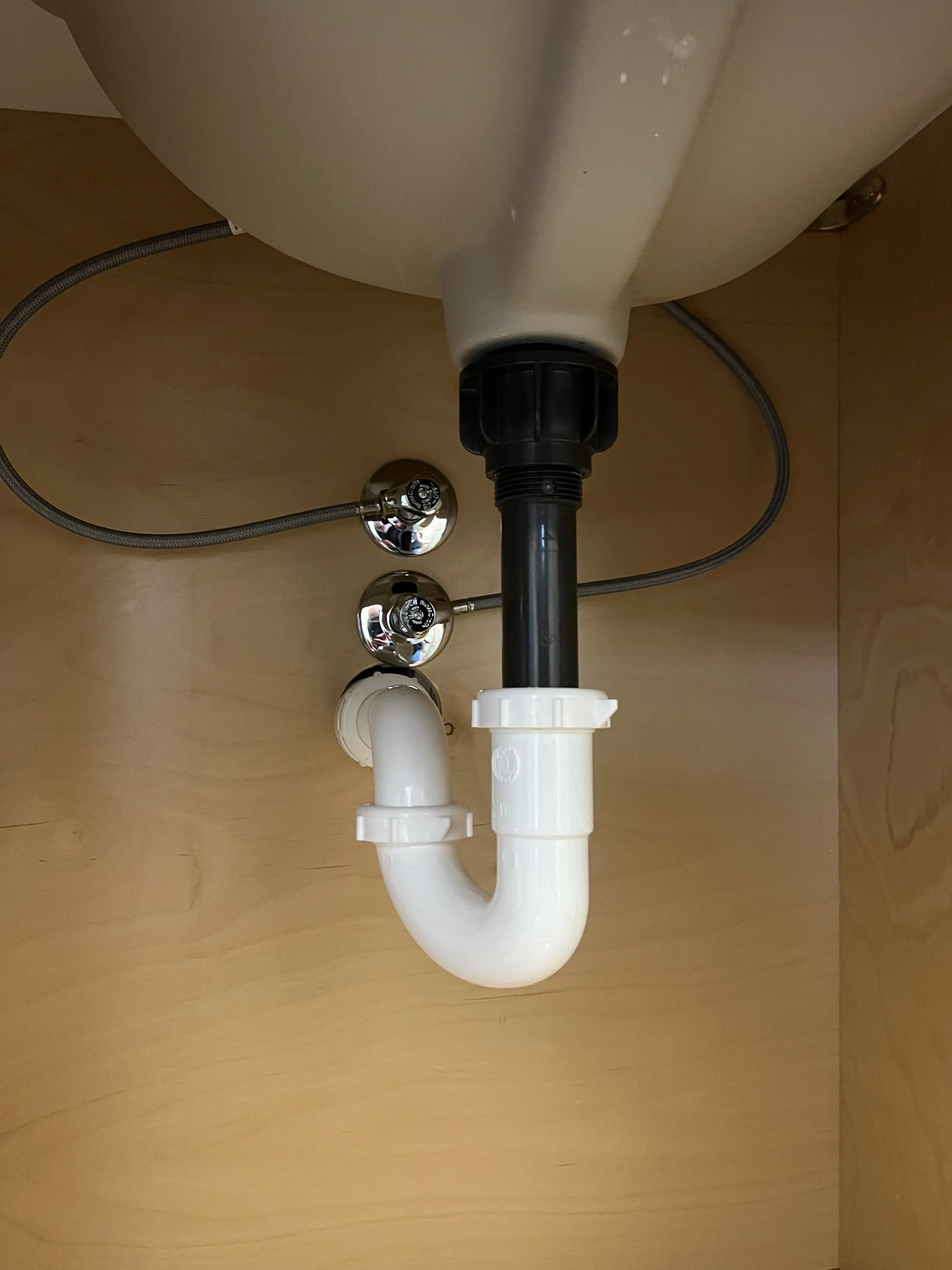 Under-sink plumbing with PVC pipes, a black waste pipe, and metal water supply lines beneath a kitchen sink