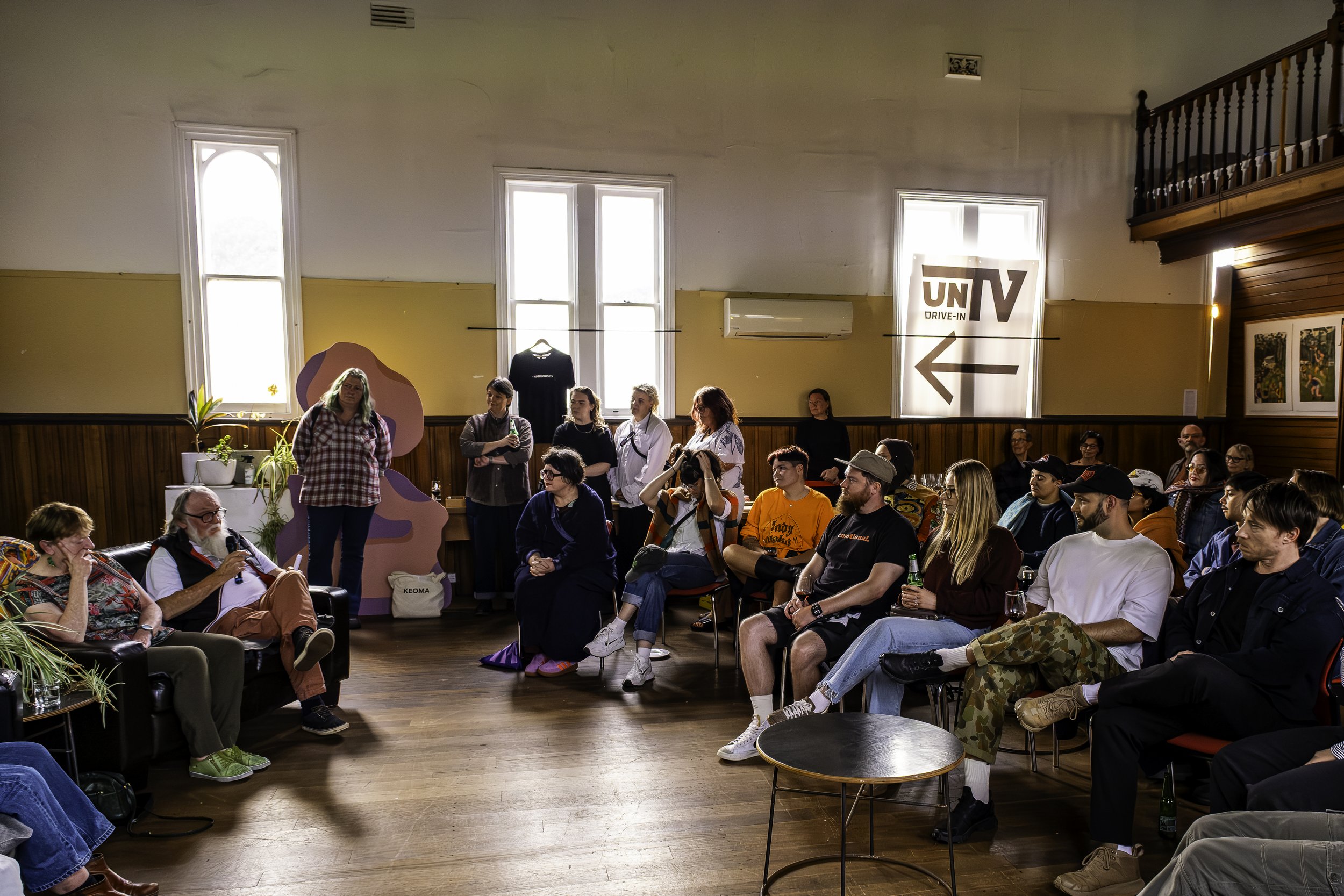 Group of people attending a discussion or presentation in a room with wooden floors and yellow walls, some seated and some standing, with a window on the back wall and a large sign with an arrow pointing left.
