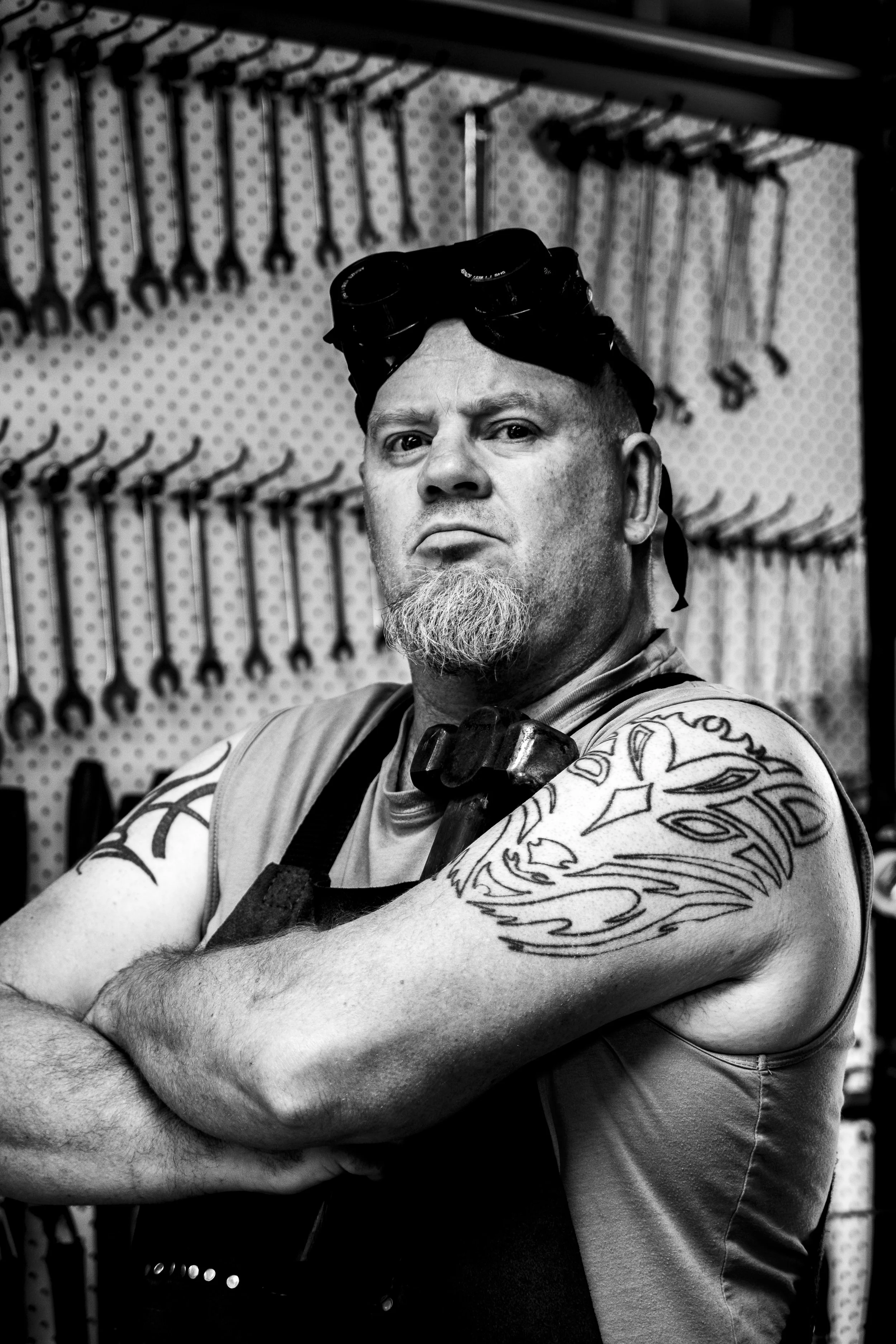 A man with tattoos on his arms, wearing a sleeveless shirt and safety goggles on his head, stands with arms crossed in front of a wall with hand tools hanging on it.