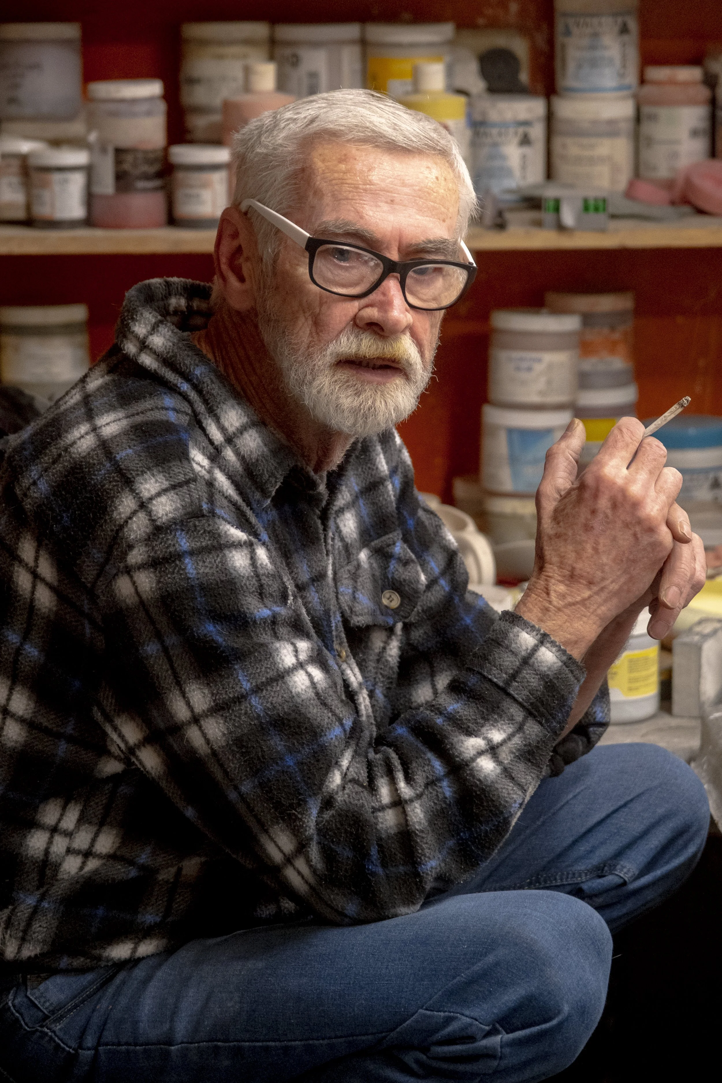 An elderly man with glasses and a beard, wearing a plaid flannel shirt, sitting in a workshop or storage area with shelves of paint containers behind him.