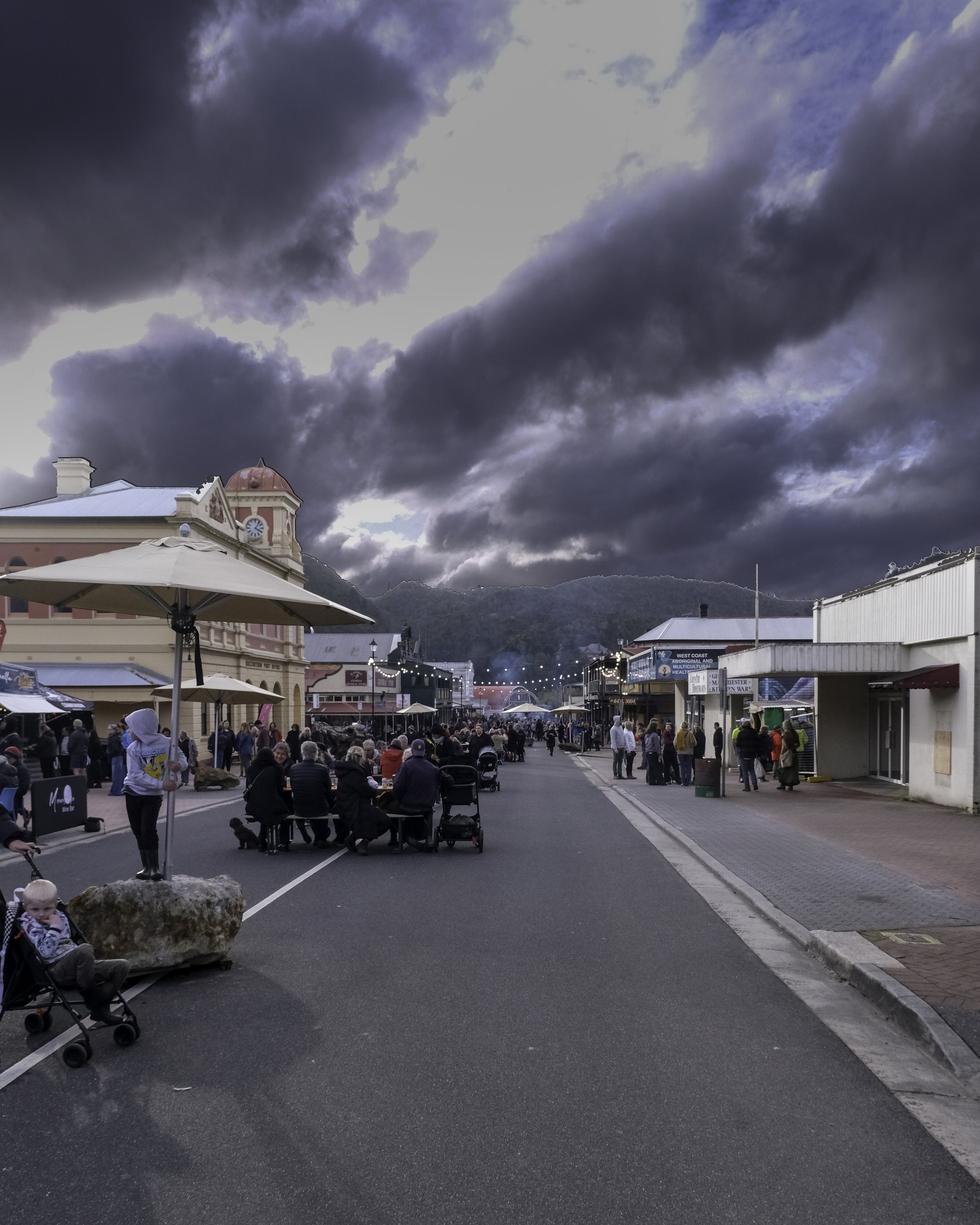 A busy outdoor street market with people sitting and walking, under cloudy skies with mountains in the background.