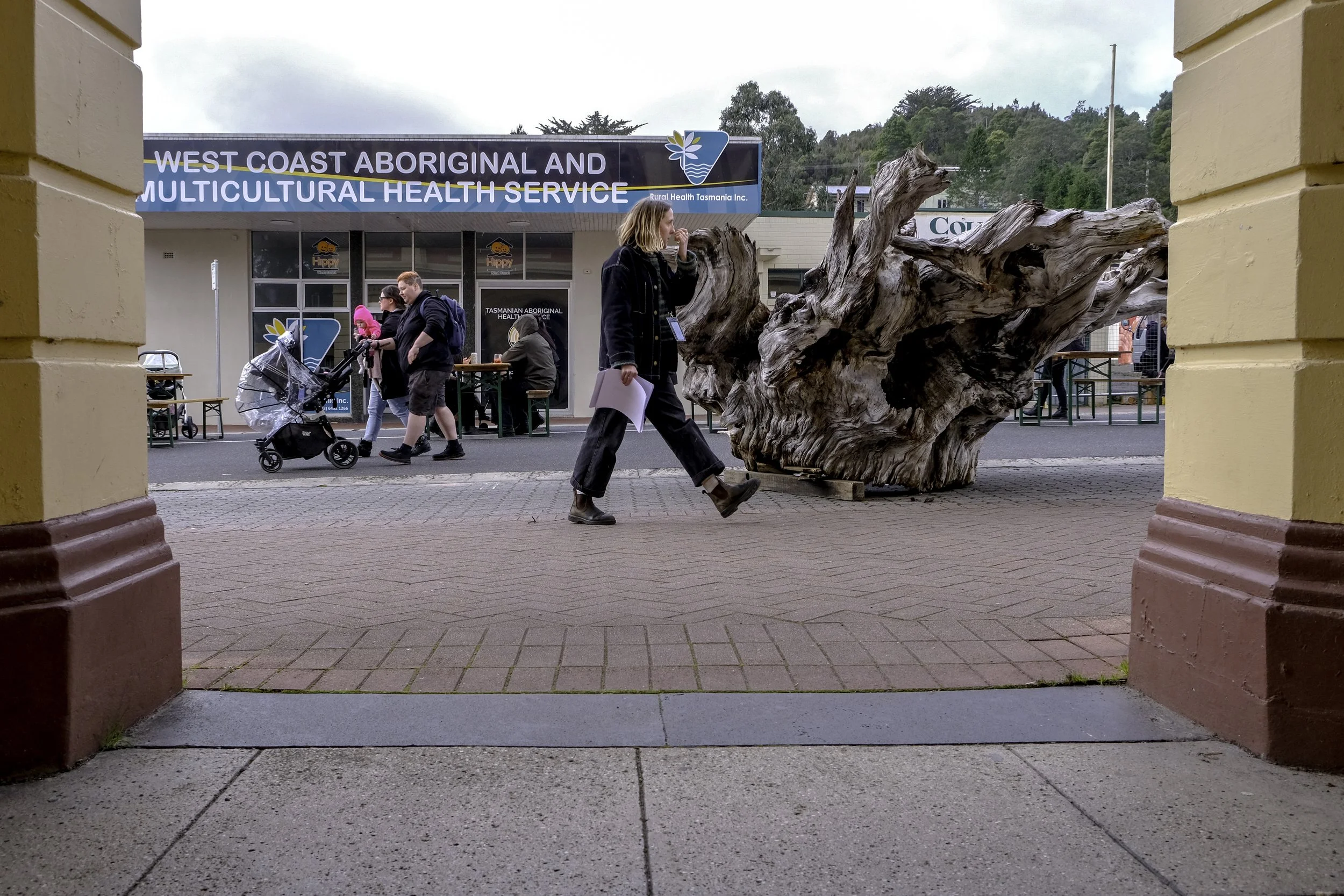 Woman walking past a large piece of driftwood outside a health services building with a sign that reads 'West Coast Aboriginal and Multicultural Health Service.' Other people are in the background, some pushing strollers and sitting at outdoor tables.