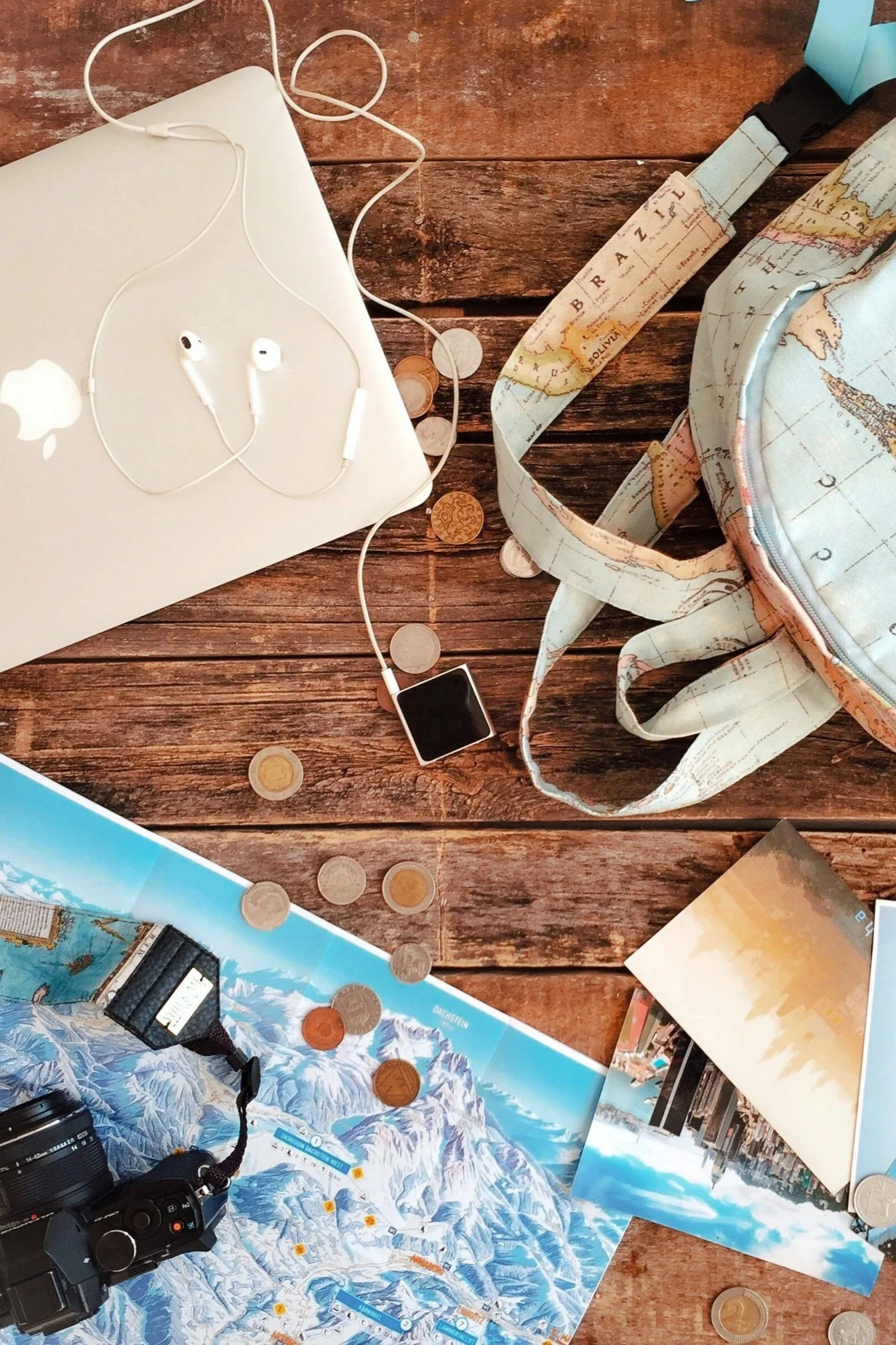 Flat lay of travel essentials on a wooden table including a laptop, camera, backpack, map, postcards, and coins