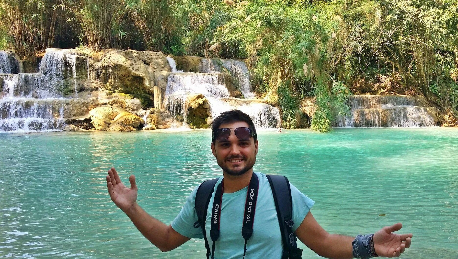 Travel advisor Kevin Miller smiling with camera at Kuang Si Waterfalls, turquoise cascading water and lush jungle in background, Luang Prabang, Laos