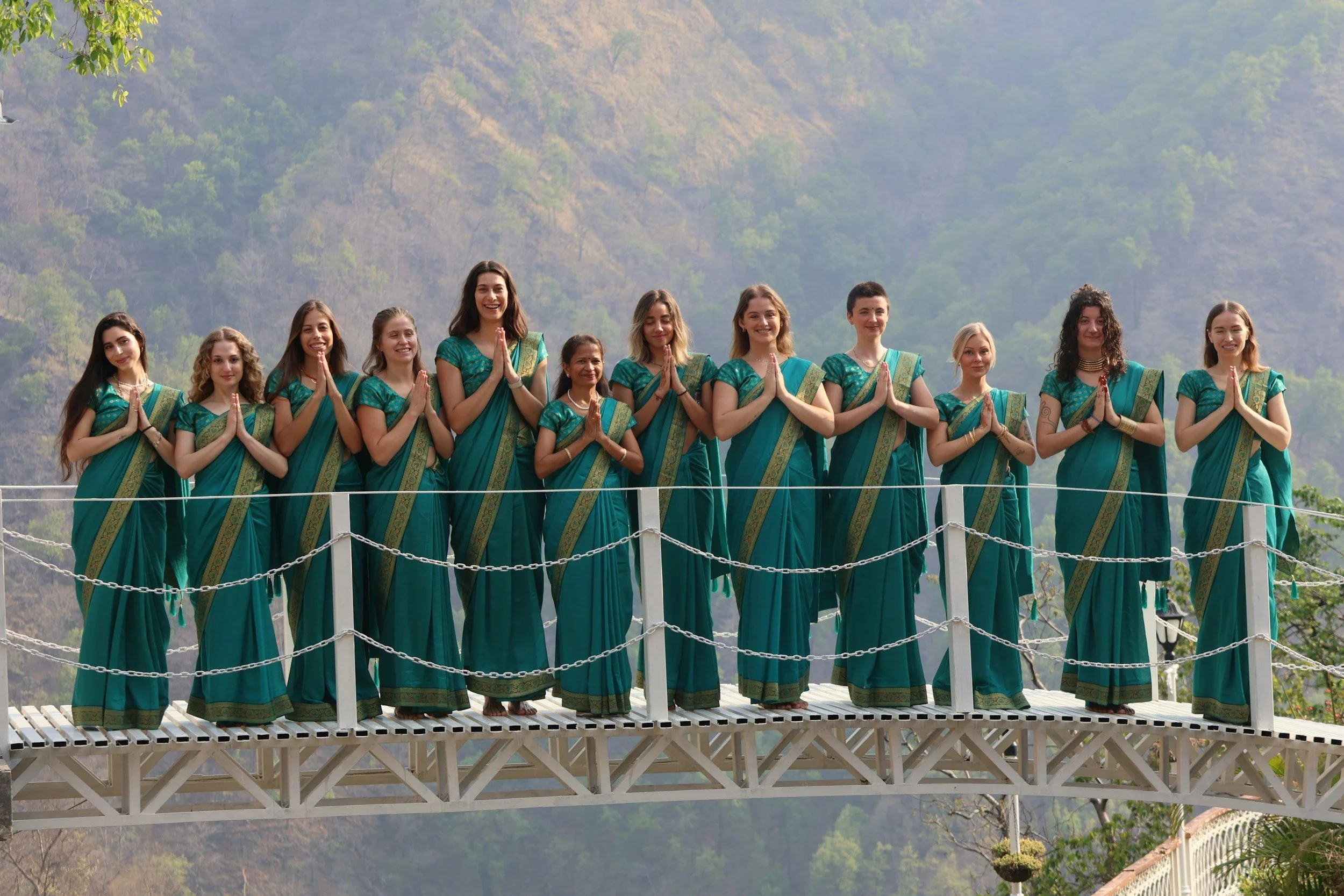 Group of twelve women in matching teal draped clothing standing on a footbridge with hands in namaste gesture, wellness retreat