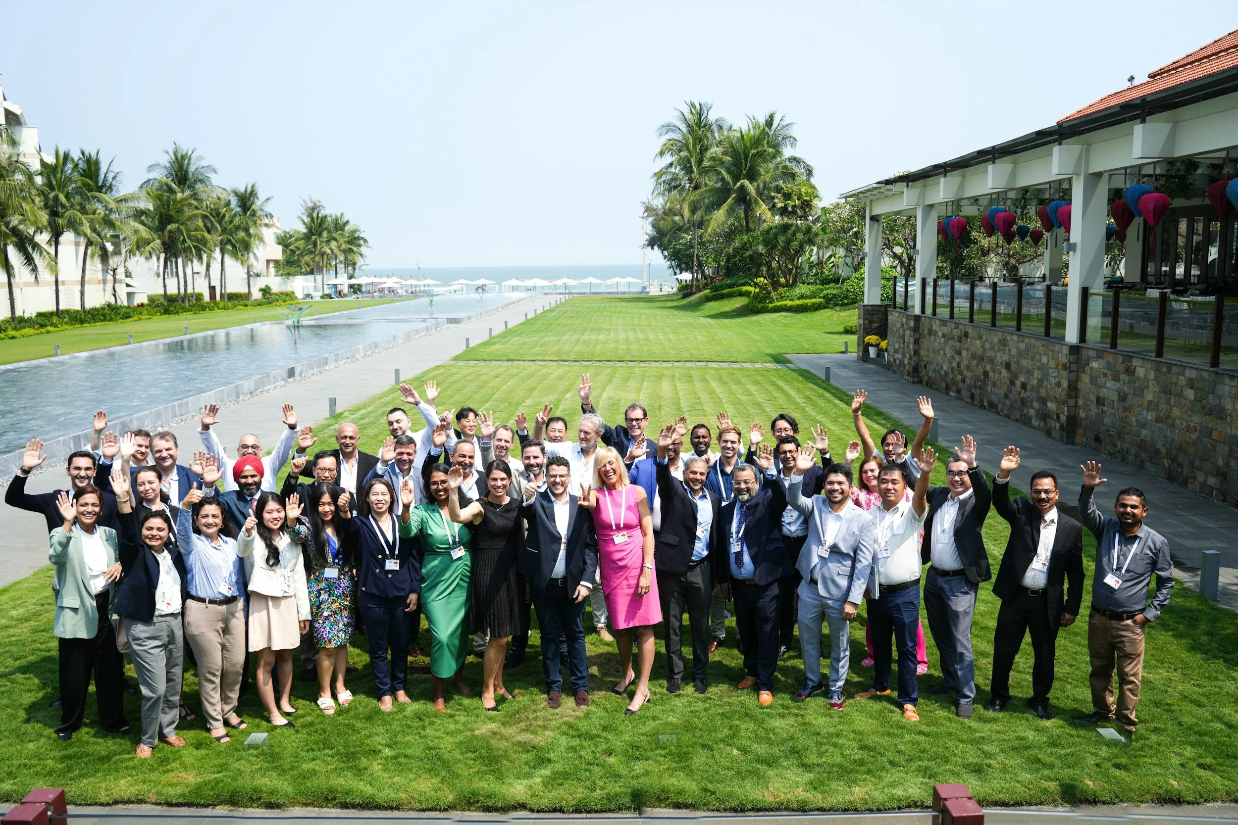 Large group of professionals in business casual attire gathered outdoors on tropical grounds with palm trees and ocean in the distance, corporate retreat