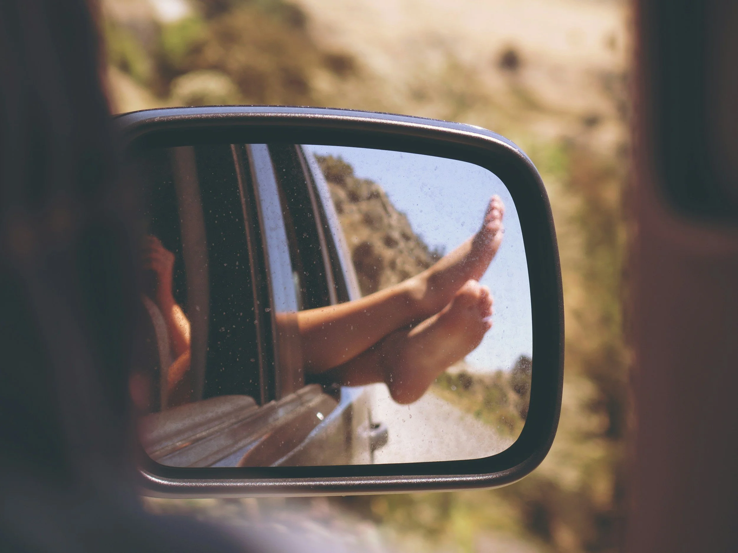 Car side-view mirror reflecting a pair of bare feet dangling out of the rear window, road trip