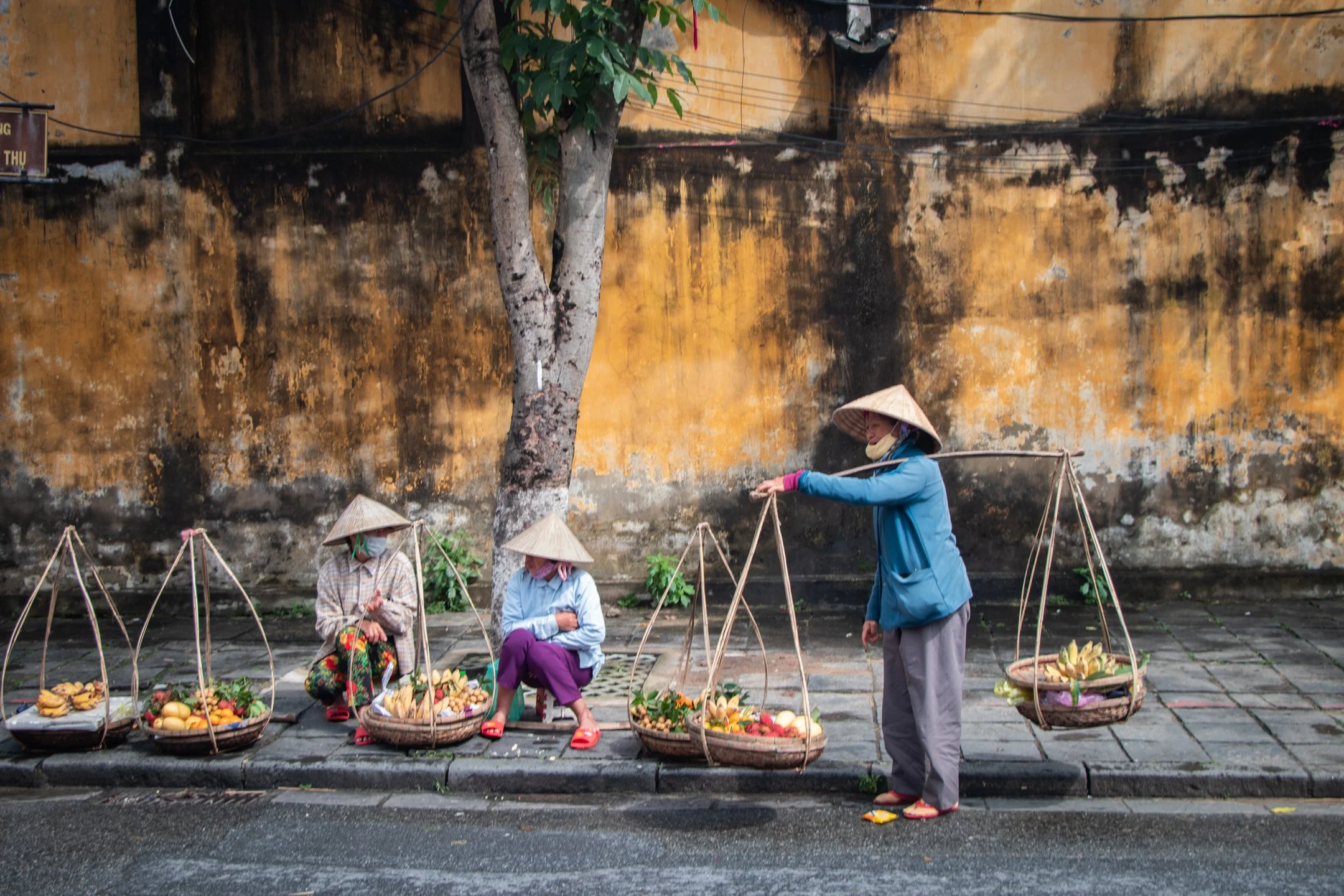 Vietnamese street vendors in traditional conical hats selling fruit from baskets against a weathered mustard-yellow wall in Hoi An's Old Town, Vietnam