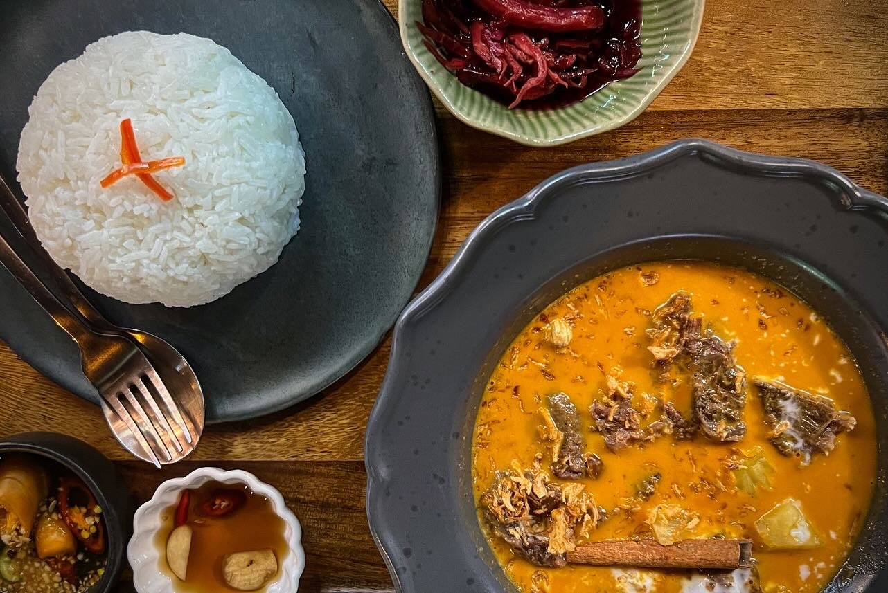 A plate of white rice garnished with orange pickled vegetables, a bowl of masaman curry with vegetables and spices, a small bowl of dipping sauce with chili peppers, and a bowl of red pickled vegetables on a wooden table.