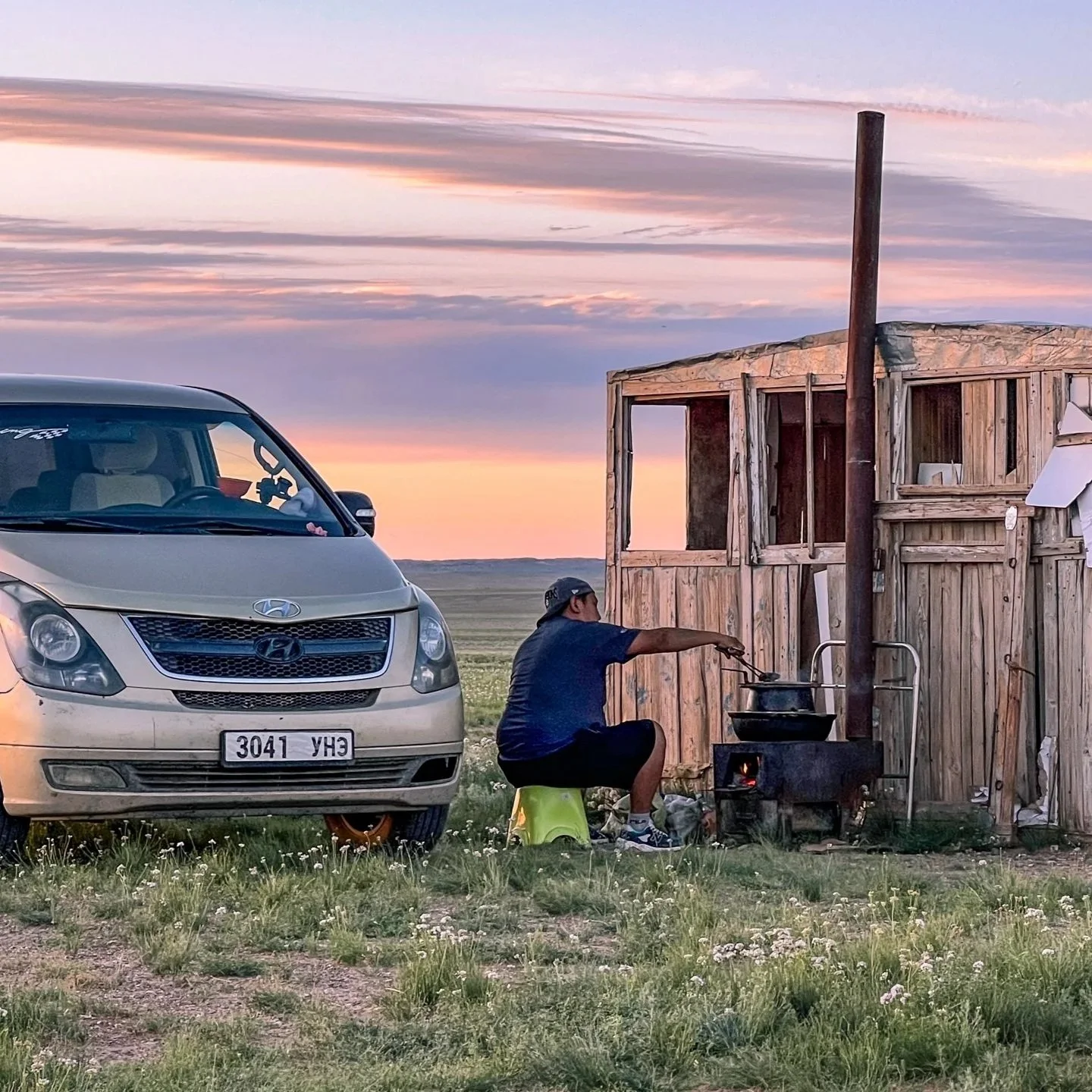 Man cooking on a traditional outdoor stove beside a van and wooden shack on the Mongolian steppe at sunset, Mongolia