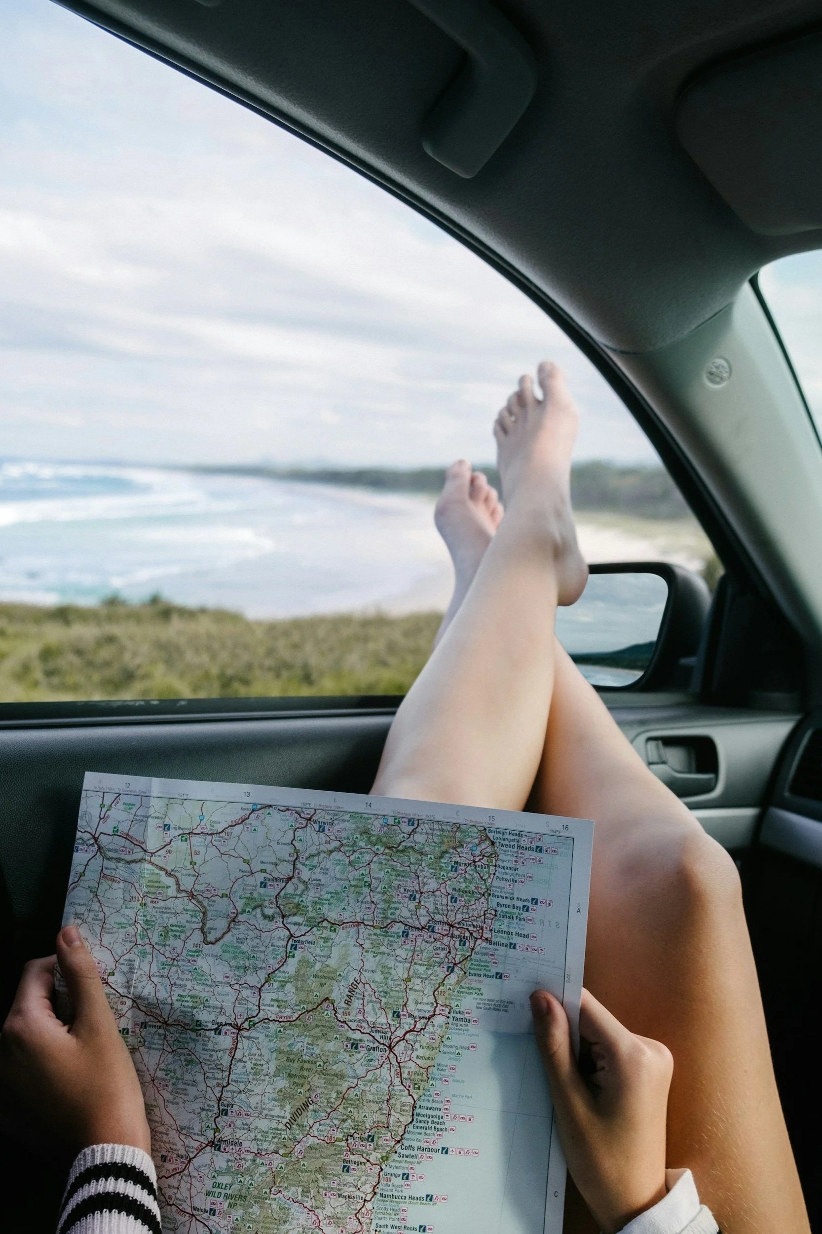 Road tripper relaxing in parked car with feet out the window holding a paper map, blurred beach in the background