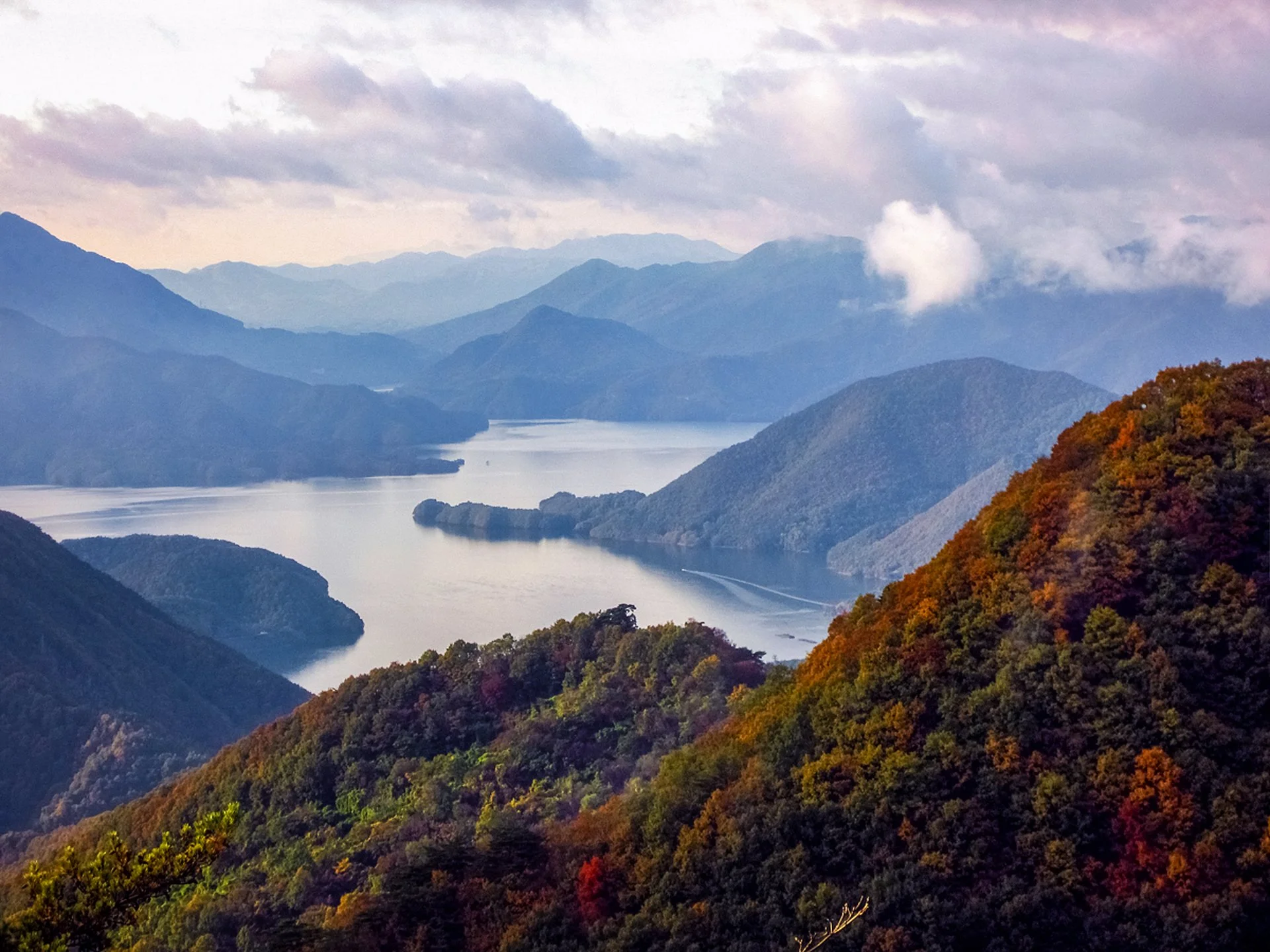 Chungju Lake reservoir surrounded by autumn foliage-covered mountains under a moody cloudy sky with soft violet hues, South Korea