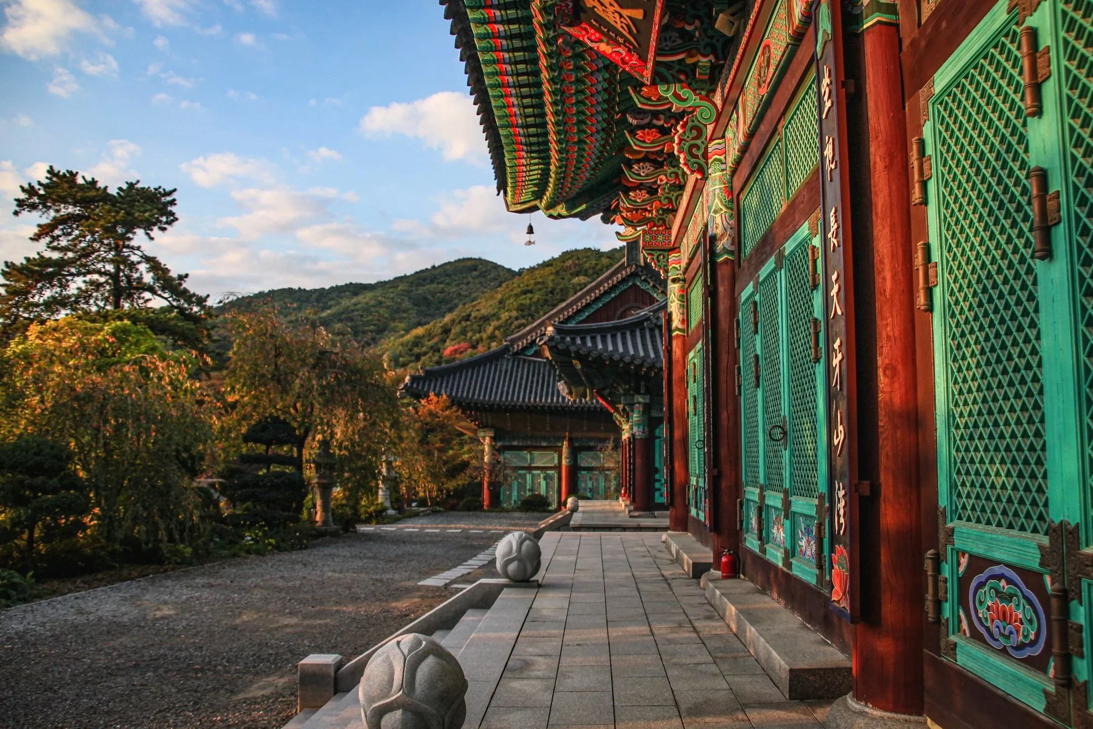 Gakwonsa Buddhist temple with ornate red and teal painted details glowing in sunset light, autumn mountains behind, Cheonan, South Korea
